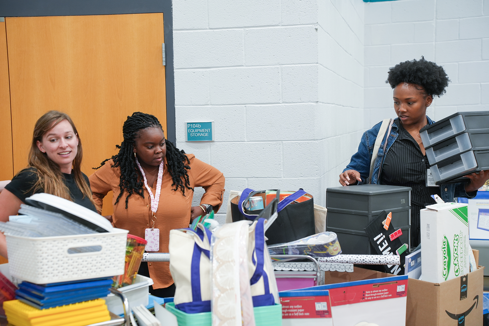 Three women stand near a table with boxes and bags, one holding a black basket, another looking at items.