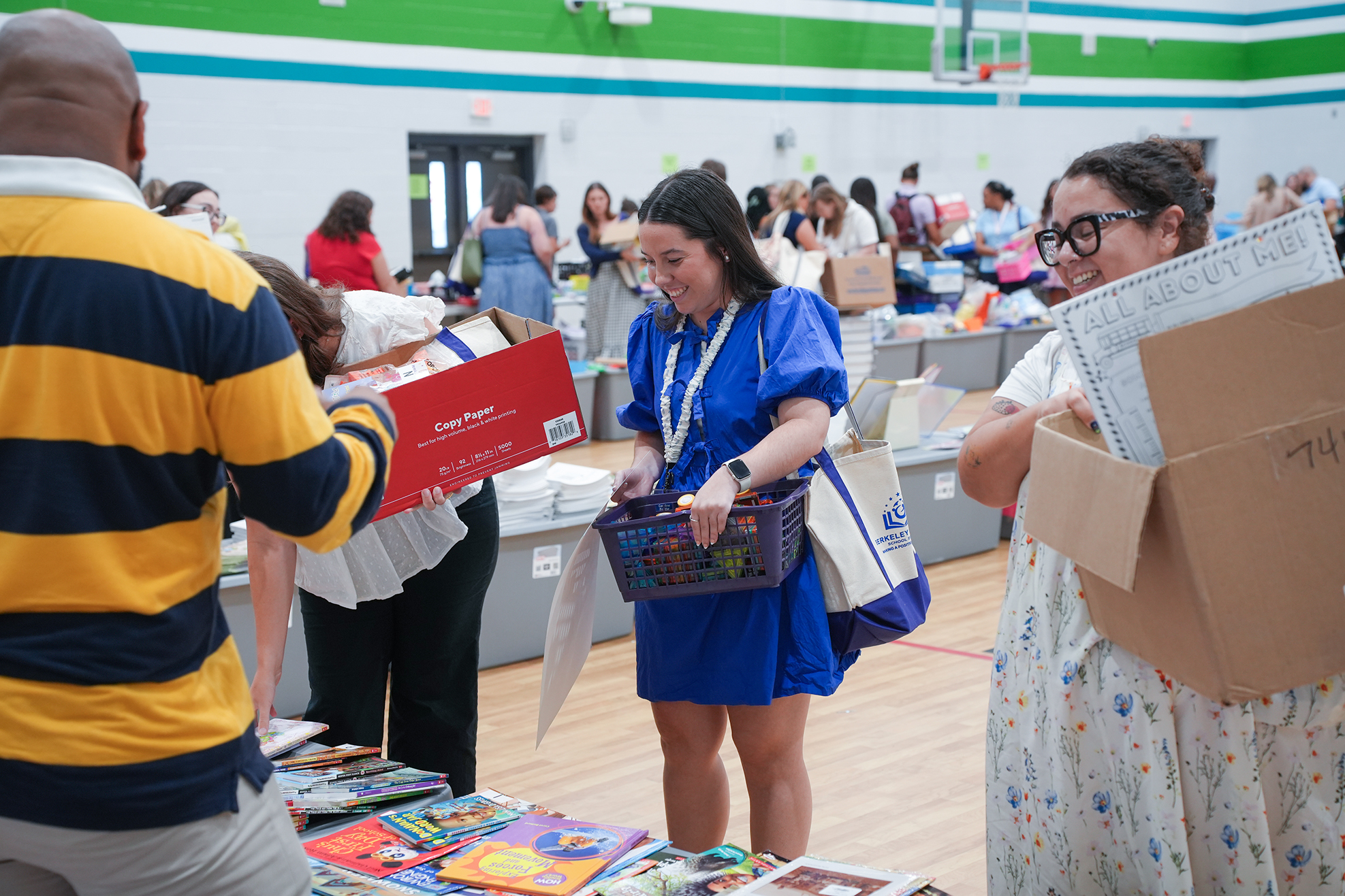 People in a gym hold boxes, books, and papers. One person hands a box to another.