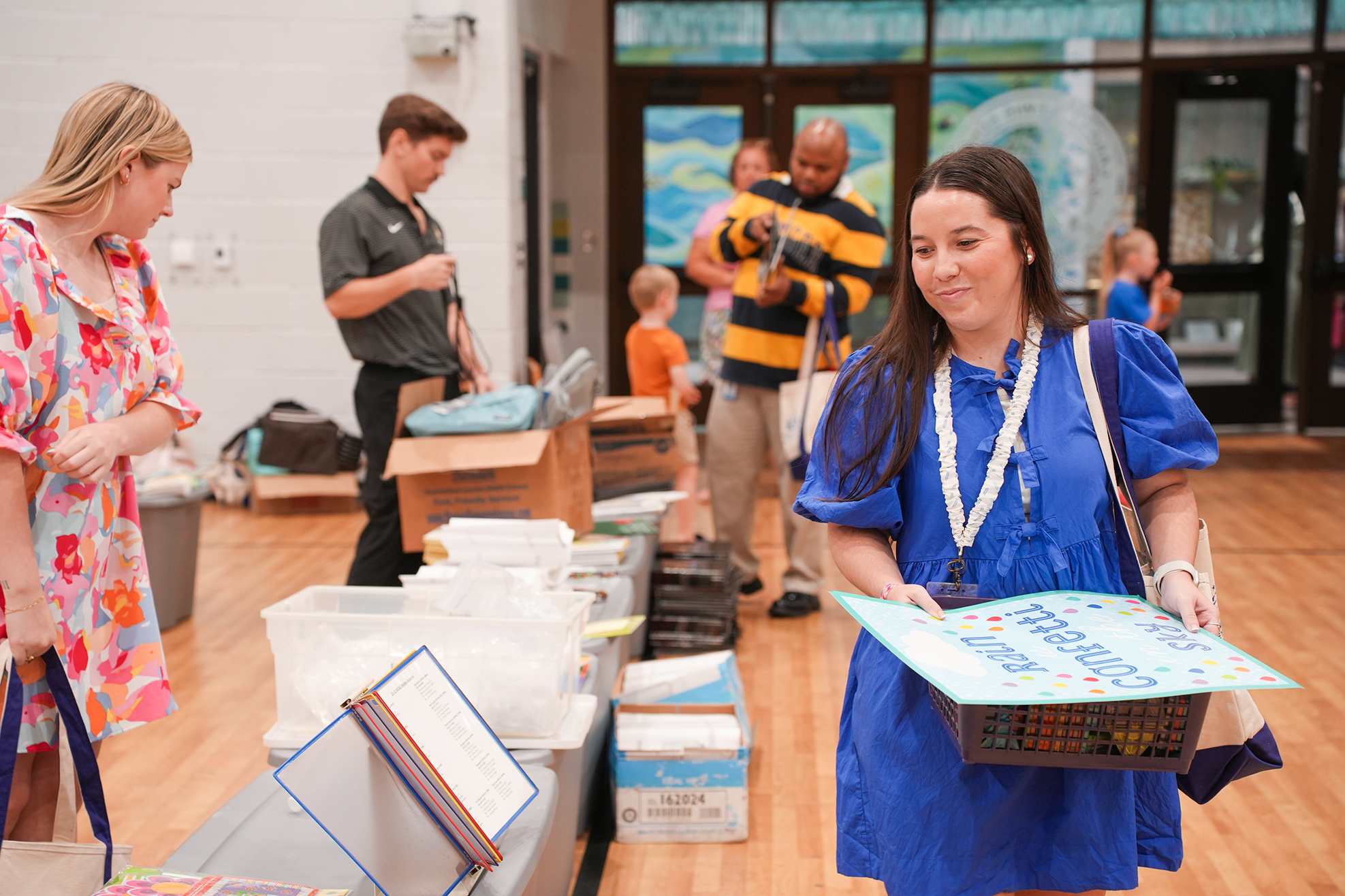 People standing in a room with boxes. One woman in blue holds a basket and paper, another woman in floral looks at her.