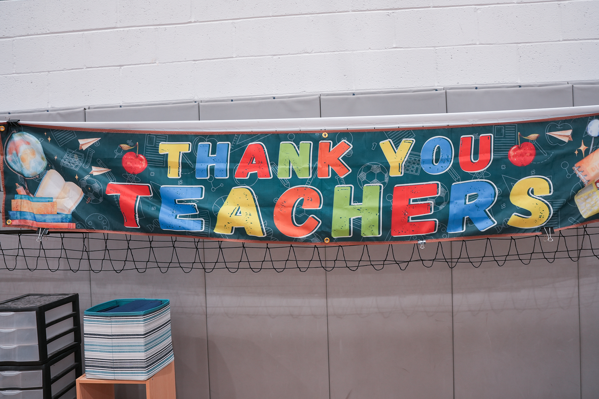 A banner saying "Thank You Teachers" in colorful letters hangs on a white wall. It includes images of books and apples.