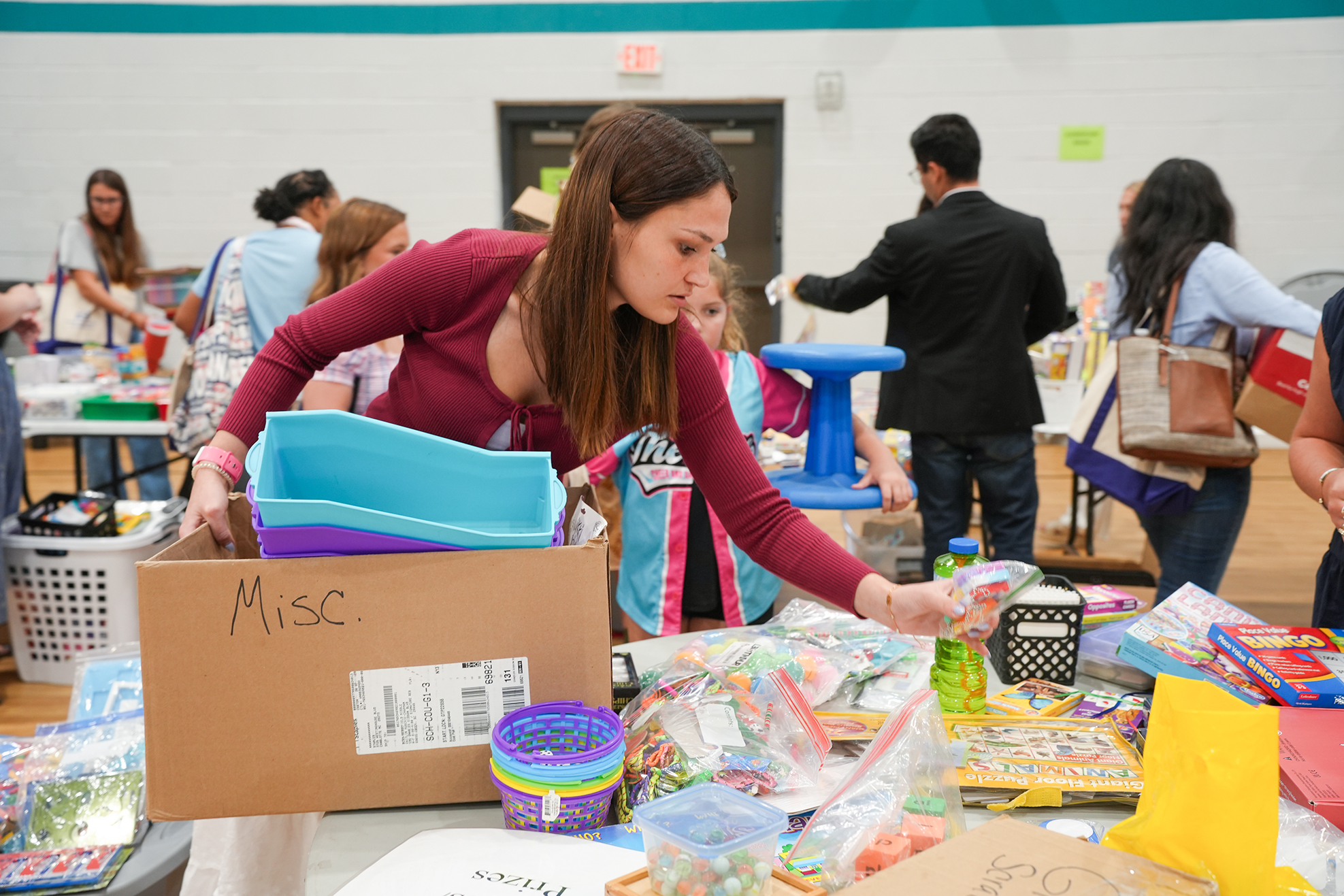 A woman in a red shirt organizes items in a box labeled "Misc" in a busy indoor area.