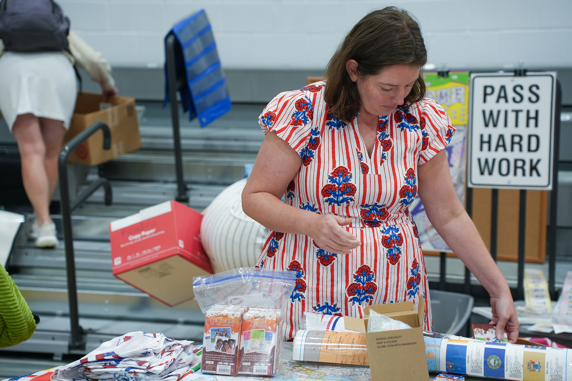 A woman in a red-and-white dress stands at a table with products and a sign.