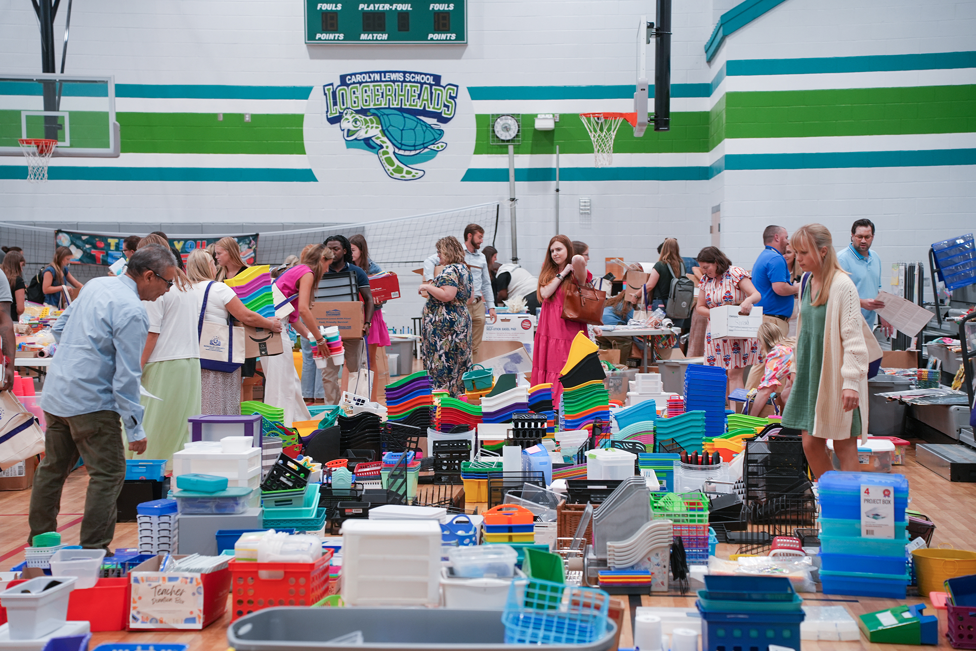 People in a gym browsing items on tables. A basketball hoop and a green and white wall with a logo.