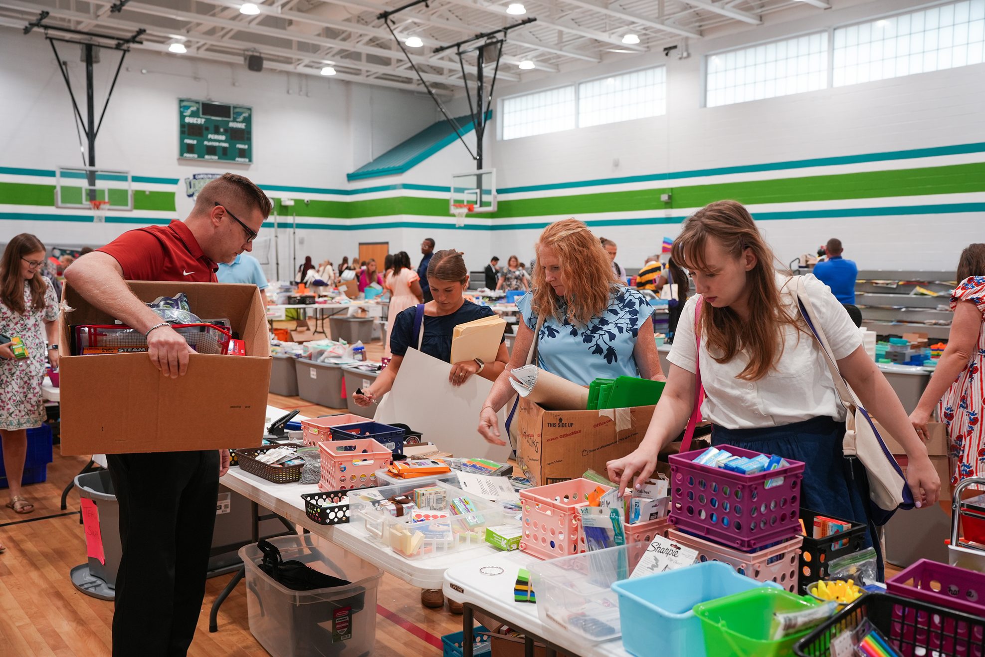 People sort through boxes in a large room, likely a gymnasium, with wooden floors and high ceilings.