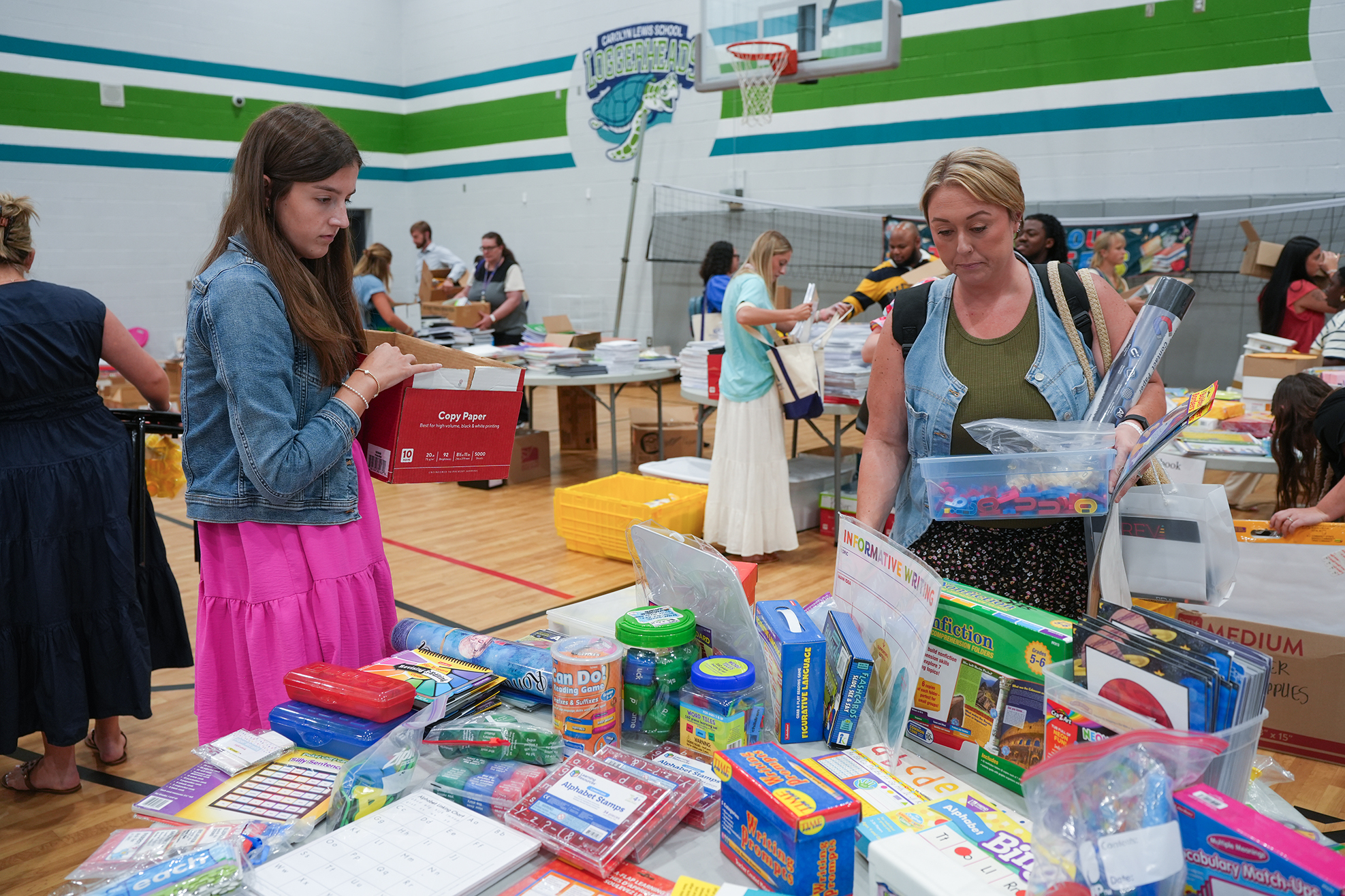 Two women examine school supplies on a table in a gymnasium. Colorful items are on display, and a basketball hoop is visible in the background.