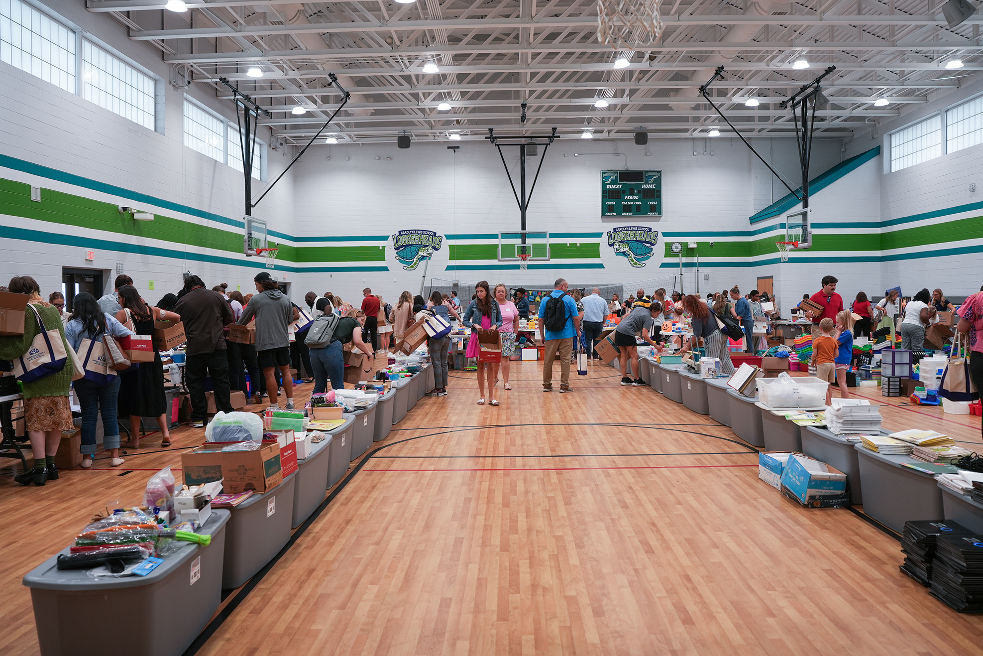 Many people gather around tables in a gymnasium, examining items. Bright lights and green and white striped walls.