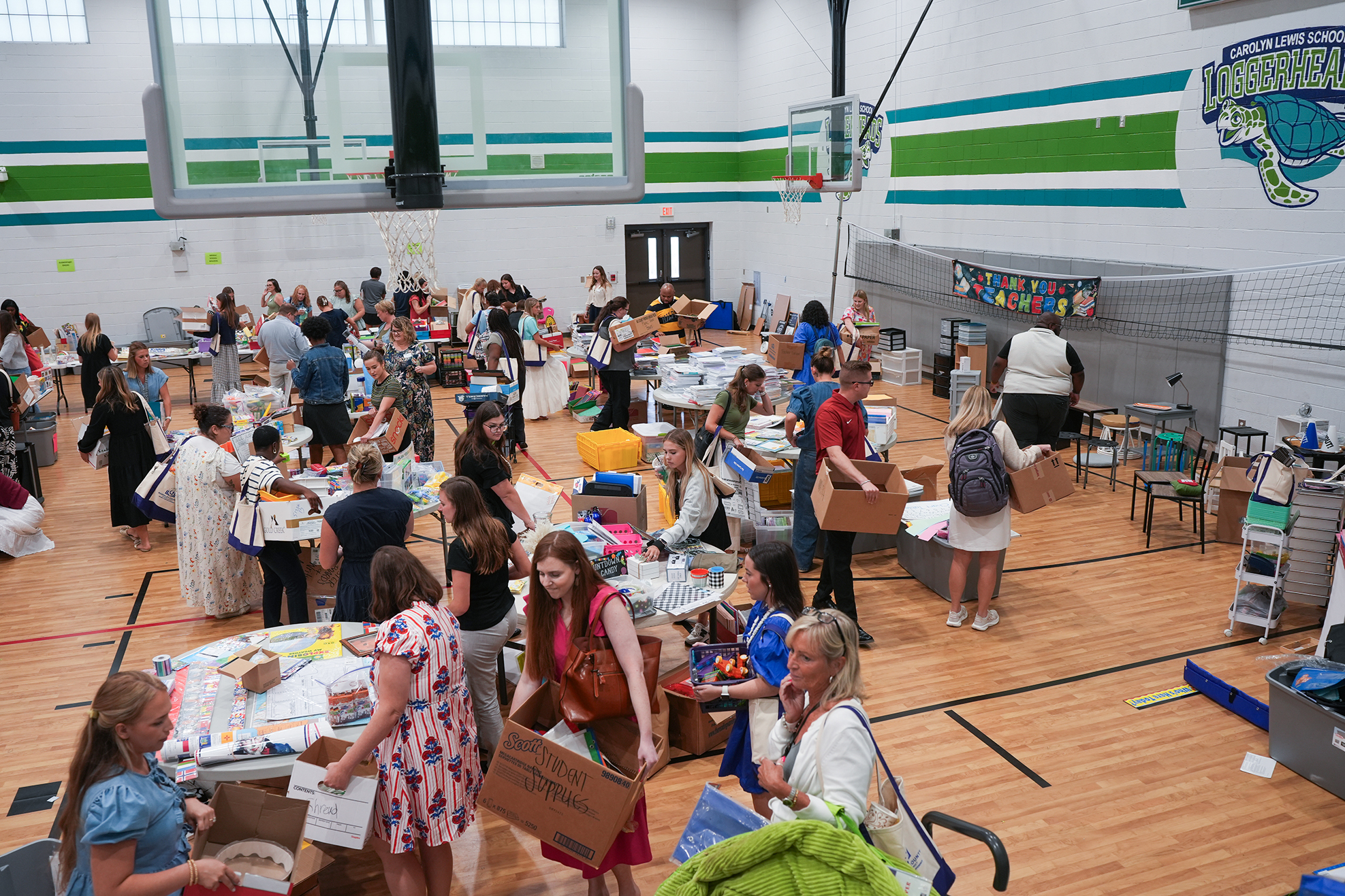 Many people in a gymnasium, engaging in activities at tables with boxes, against a backdrop of a basketball court.