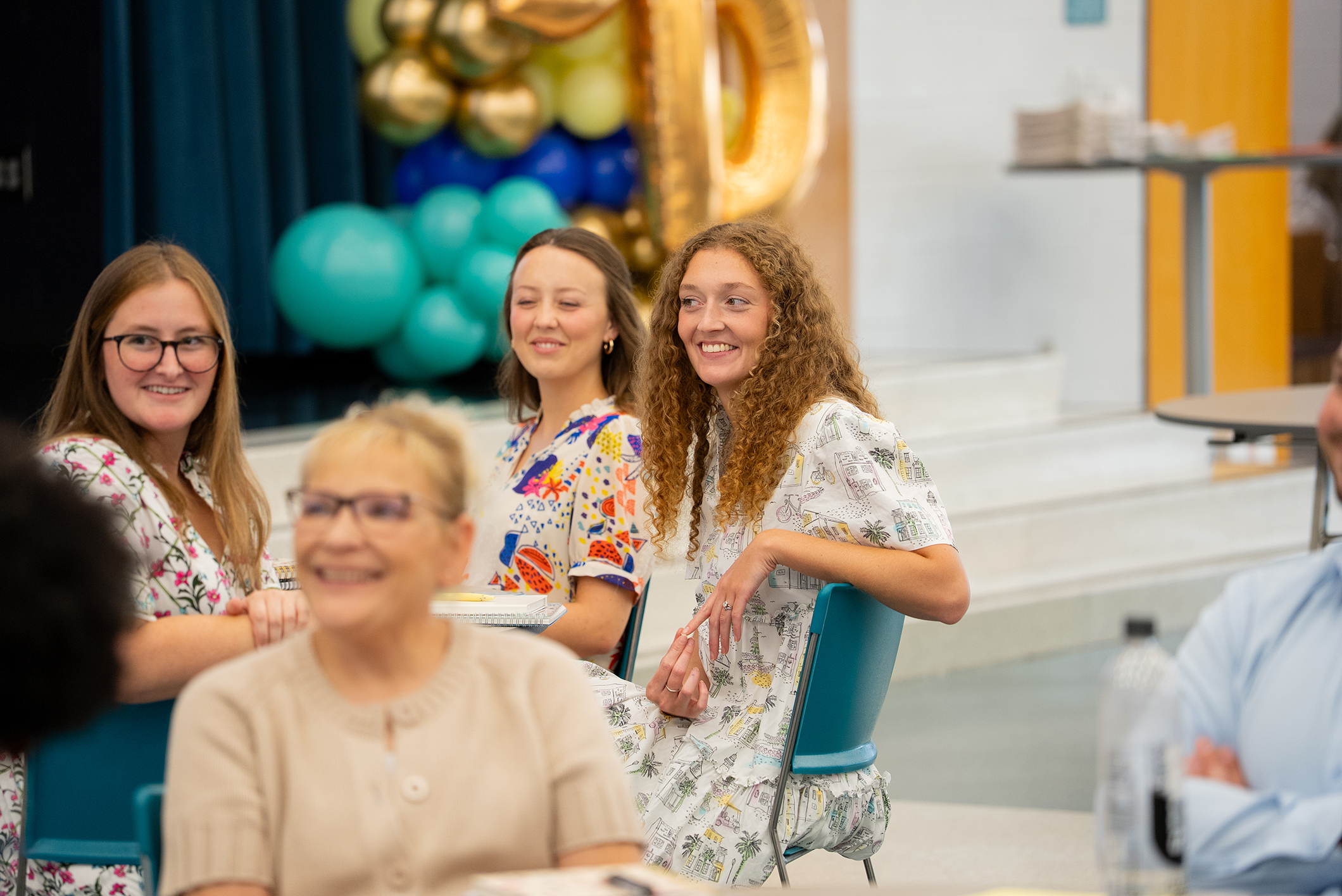 Four women in light dresses smile while sitting around a table in a room with balloons and a gold number.