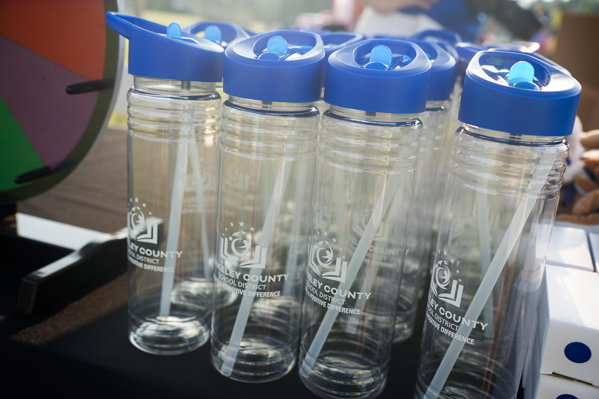 Five clear water bottles with blue lids and straws in a row on a table.