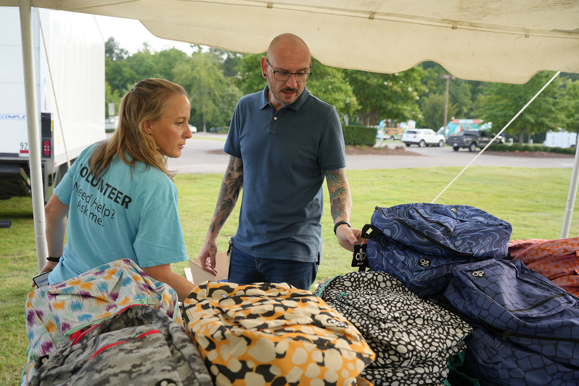 A man and a woman are standing under a tent. The man holds a bag and the woman inspects the bags on the table.