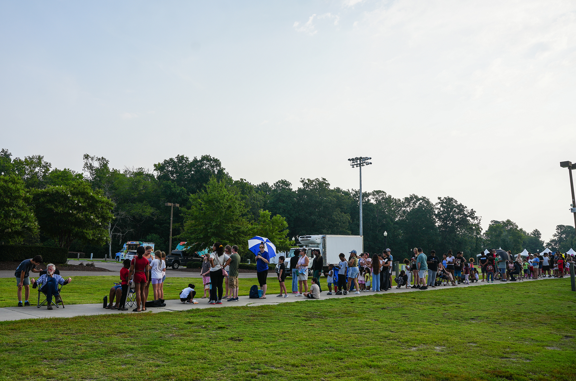 Long line of people standing on a grassy pathway with trees and a street light in the background.