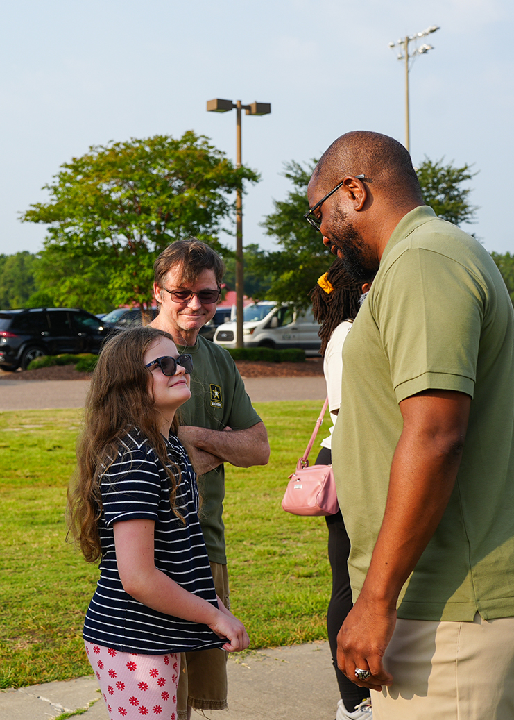 Four people on a sidewalk. A man, woman, and girl, and a woman with a pink purse. Background includes parking lot.