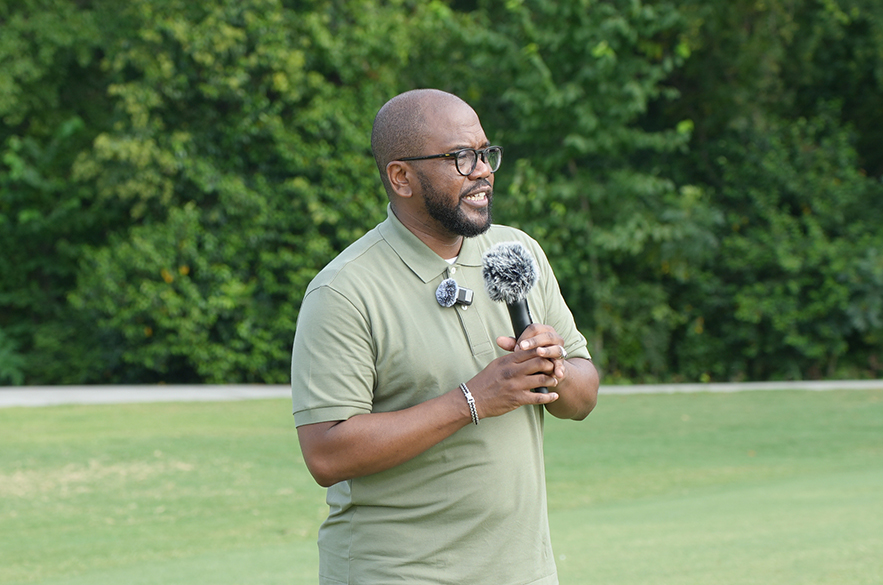 A man with glasses stands in a green field, holding a microphone, with trees in the background.