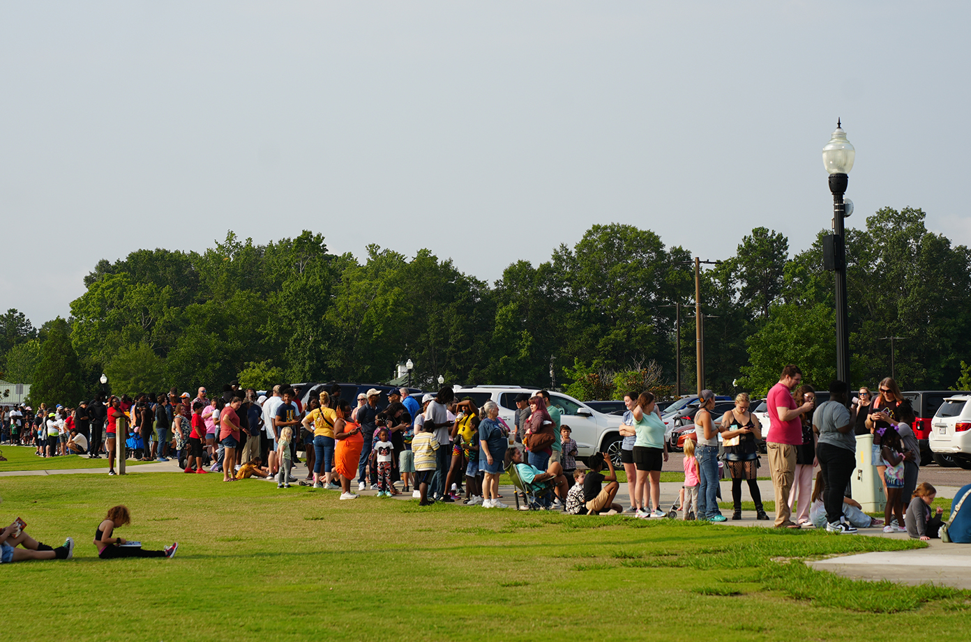 People standing in a line on a grassy field, some wearing sneakers, in a park with trees and parked cars.