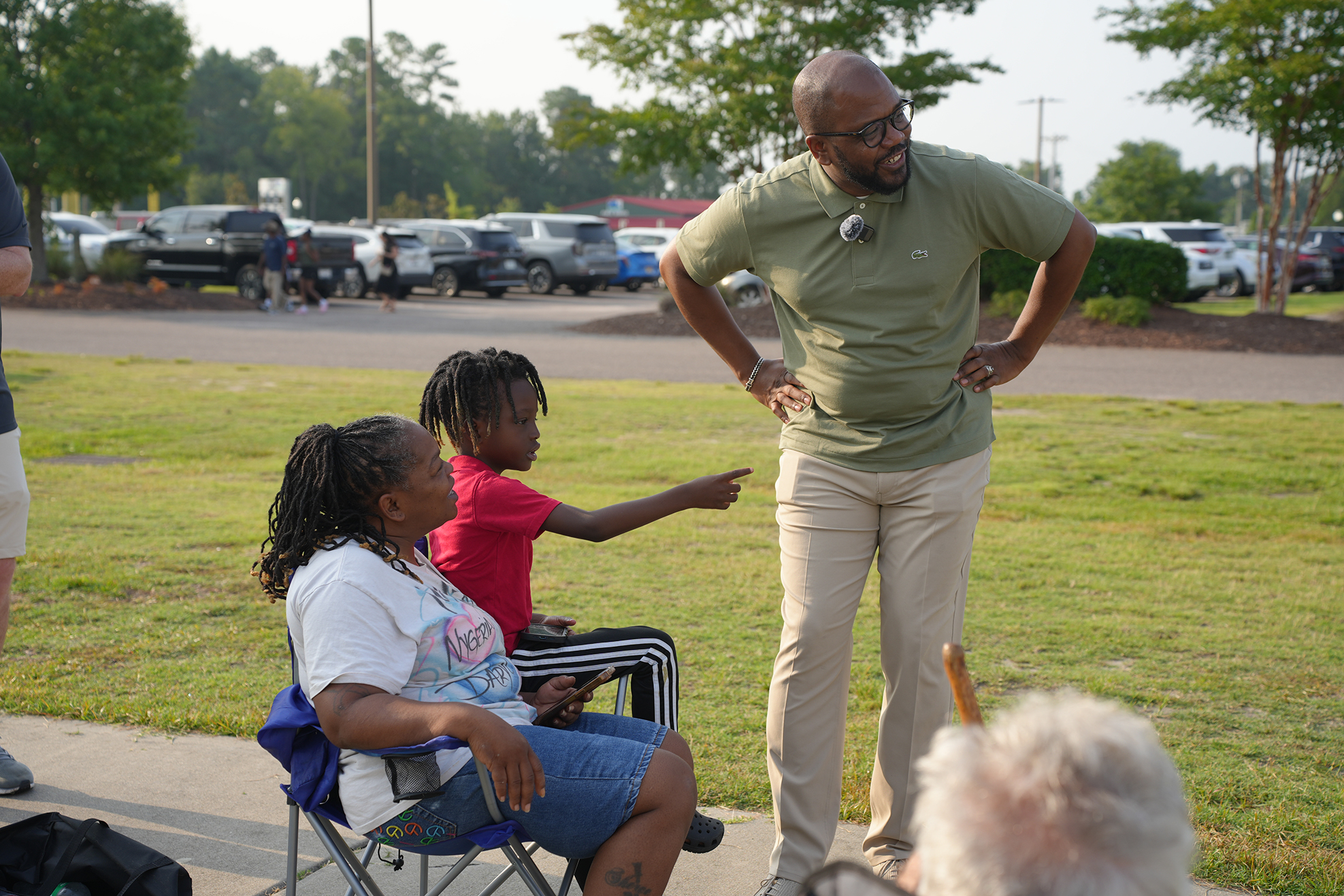 An adult man and woman speak to a group; a woman sits in a chair and a girl sits behind her.