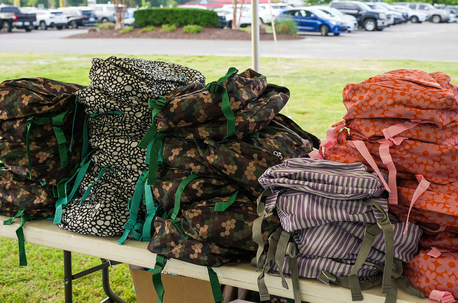 Stacks of backpacks on a table outdoors with cars parked in a lot in the background.