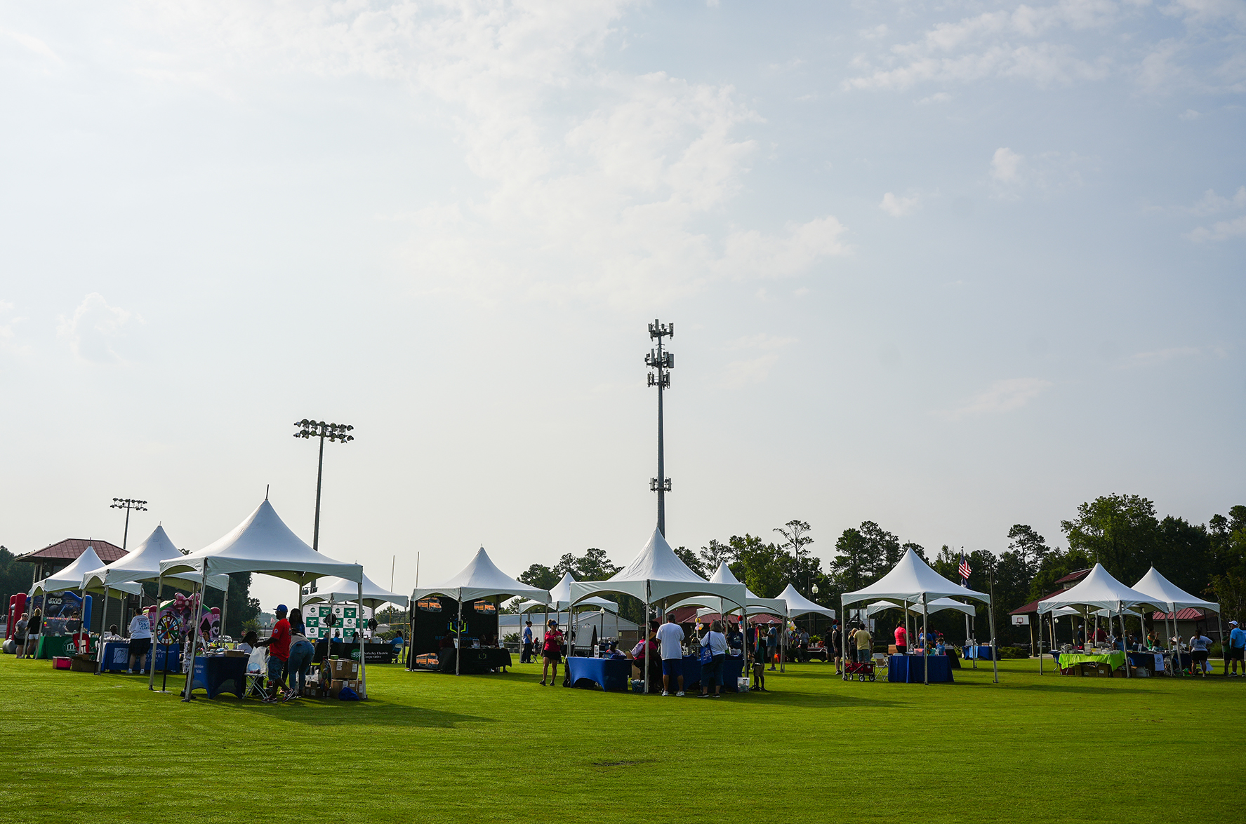 A field with several white tents set up, and people are gathered around. The sky is partly cloudy.