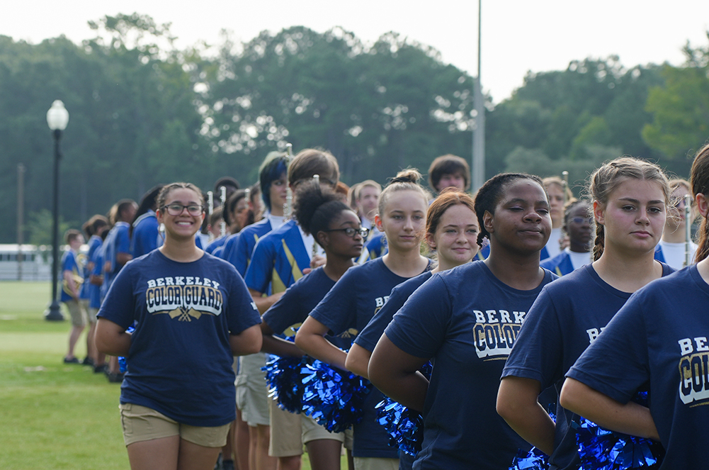 A group of people in blue shirts with white lettering stand on a grassy field, with trees in the background.