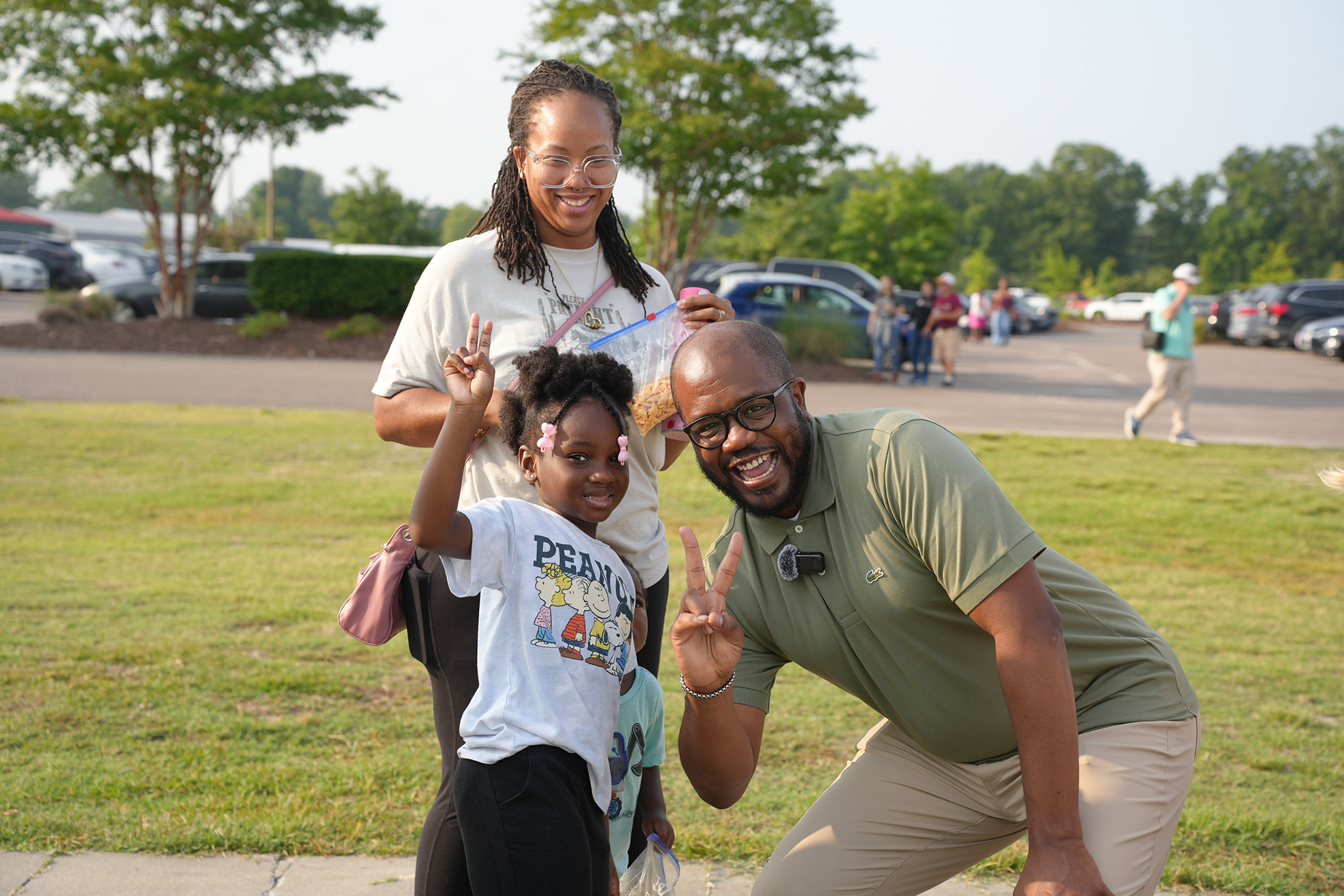 Three individuals pose in a park, with a person crouching and making peace signs, and a girl smiling.