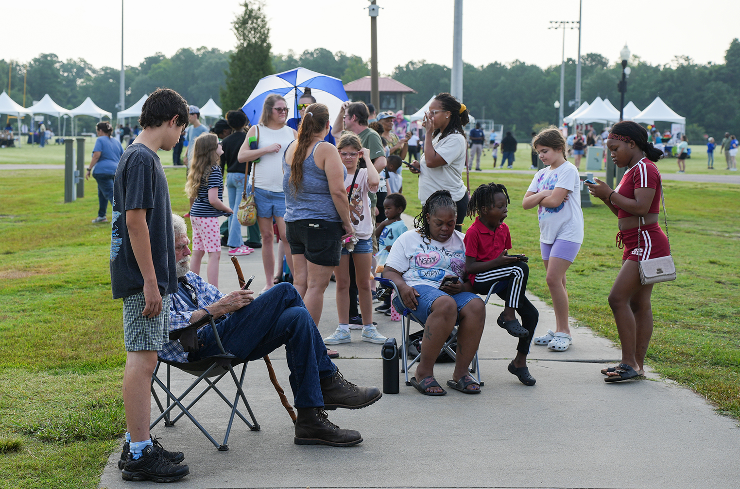Group of people at an outdoor event. Some sit on chairs on a walkway. A man sits with a cane.