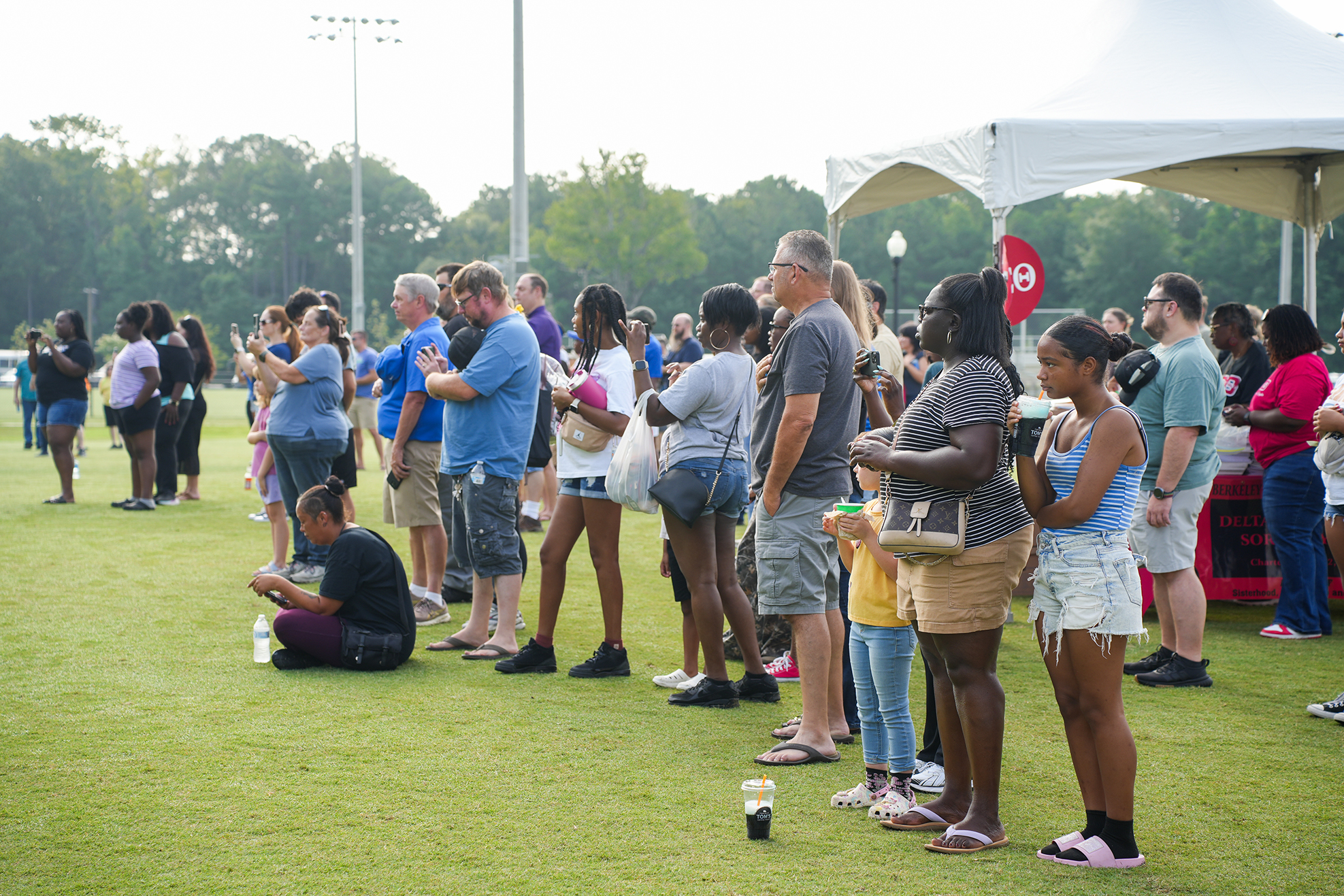 A group of people wait in line under a tent on a grass field. Some wear sandals, while others wear sneakers.
