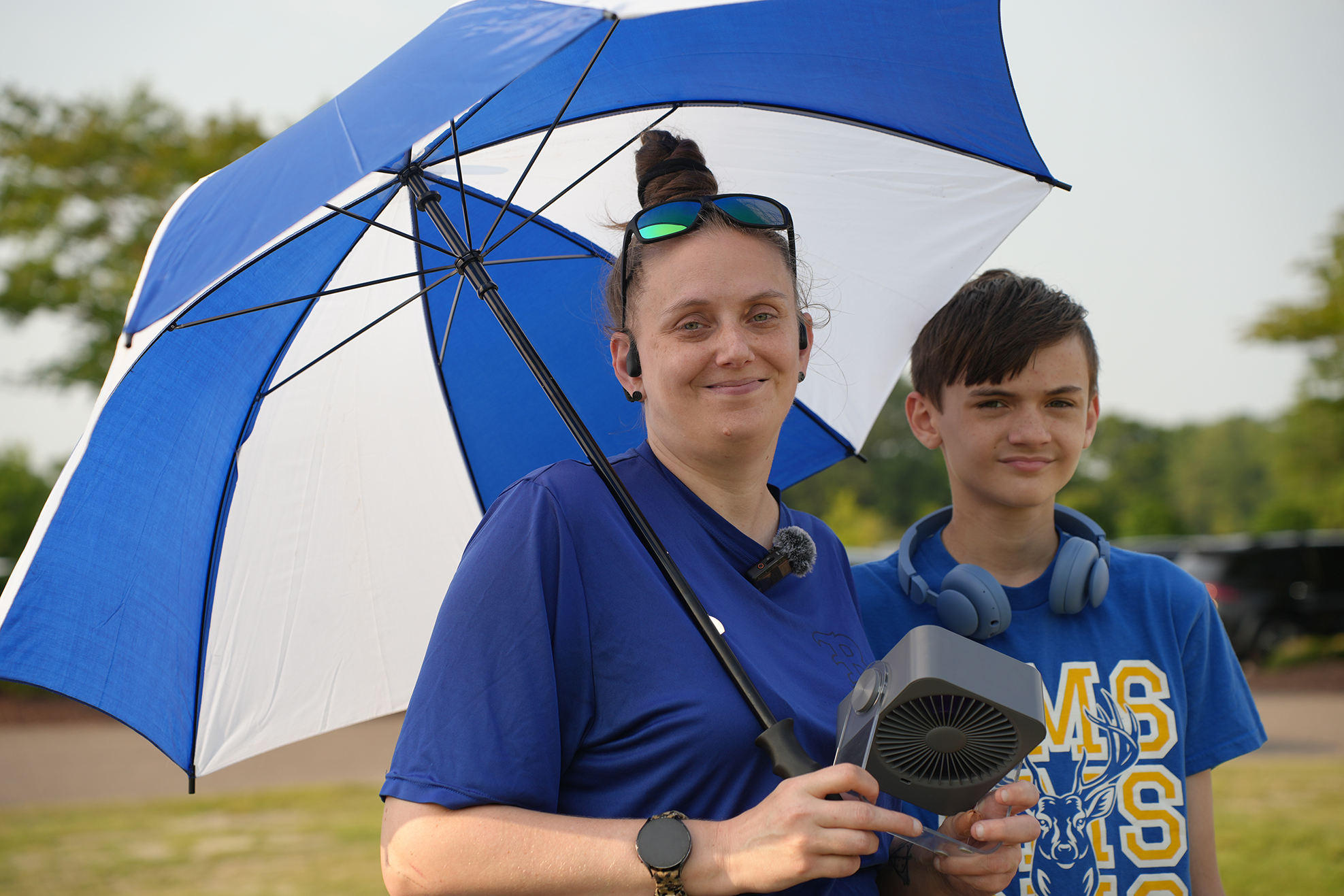 Two people, a woman with sunglasses on her head and a boy, hold a device. She holds an umbrella.