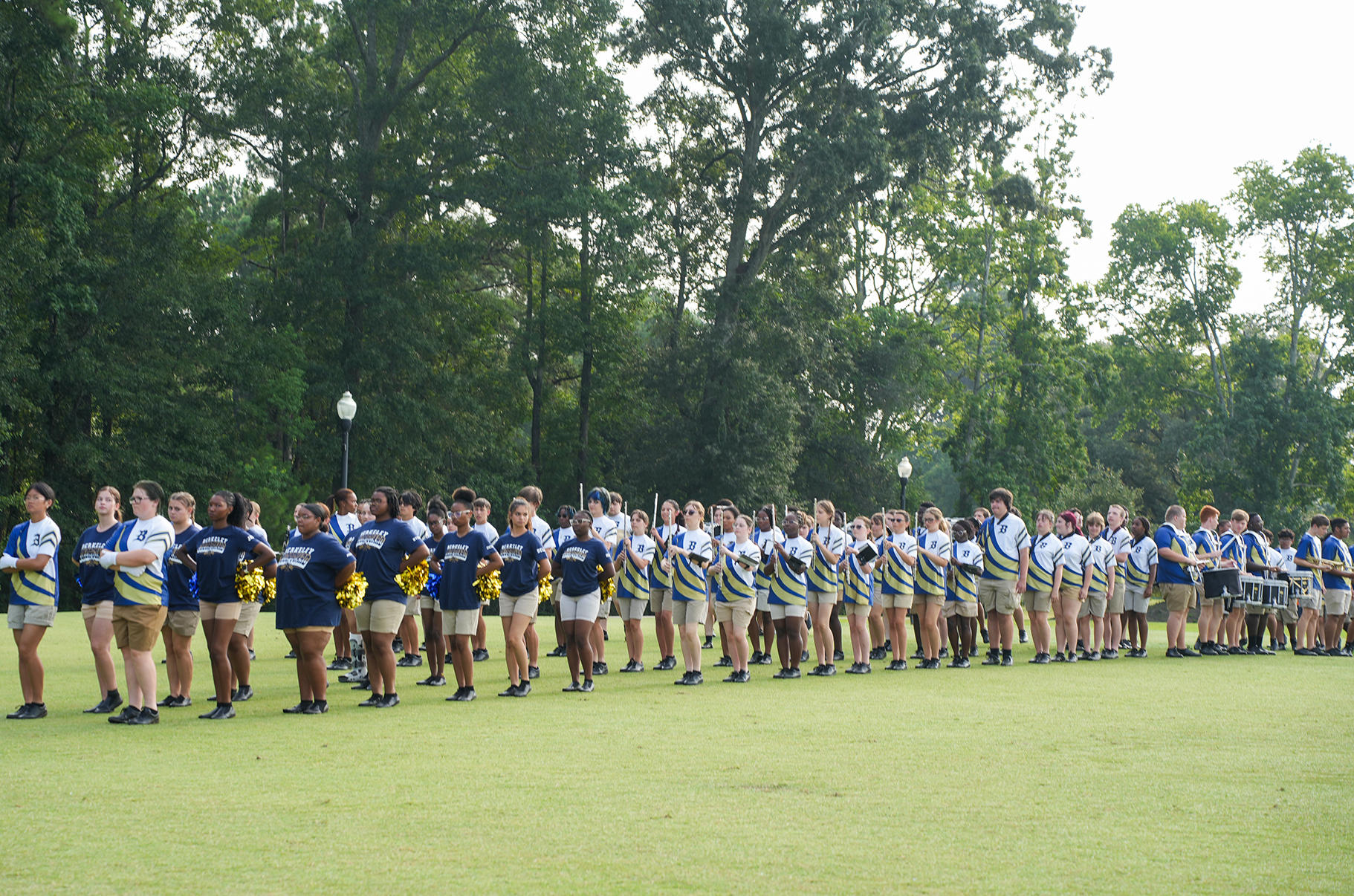 A group of people in blue, white, and yellow uniforms lined up on a grassy field with trees.