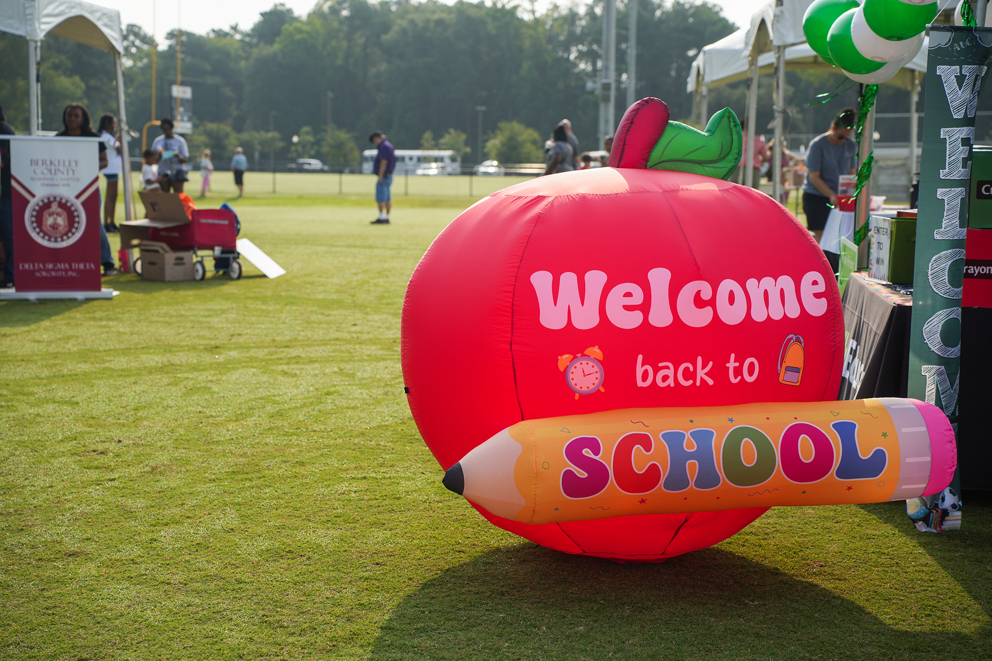 Red apple shaped balloon with "Welcome back to SCHOOL" on green grass with tents, tables, and people.