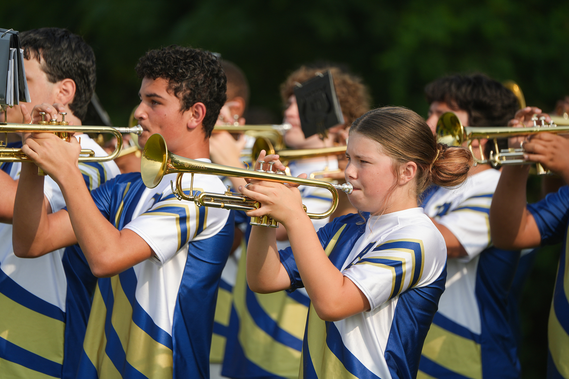 A group of young musicians in blue and white uniforms play trumpets in an outdoor setting.