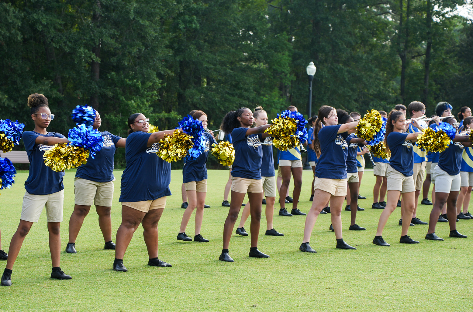 A group of cheerleaders in blue tops and khaki shorts perform on a green lawn with trees in the background.