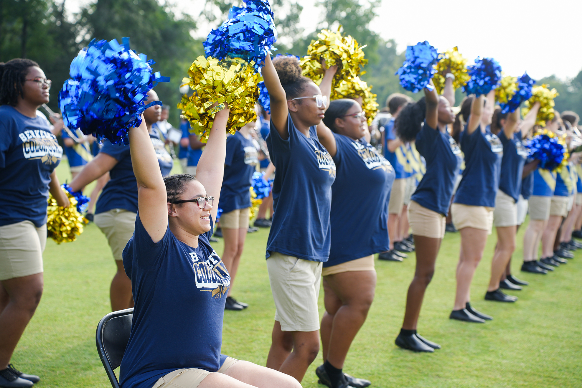Group of women with blue and yellow pom-poms standing in a line on a grassy field.