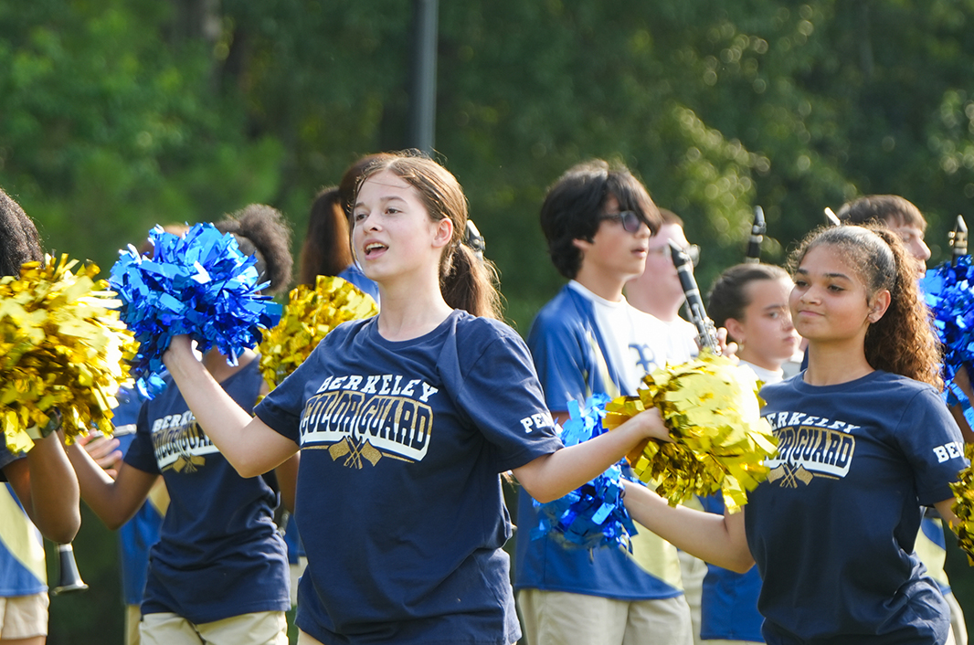 Several children in navy blue shirts hold pom-poms and clap. Behind them, others hold musical instruments.