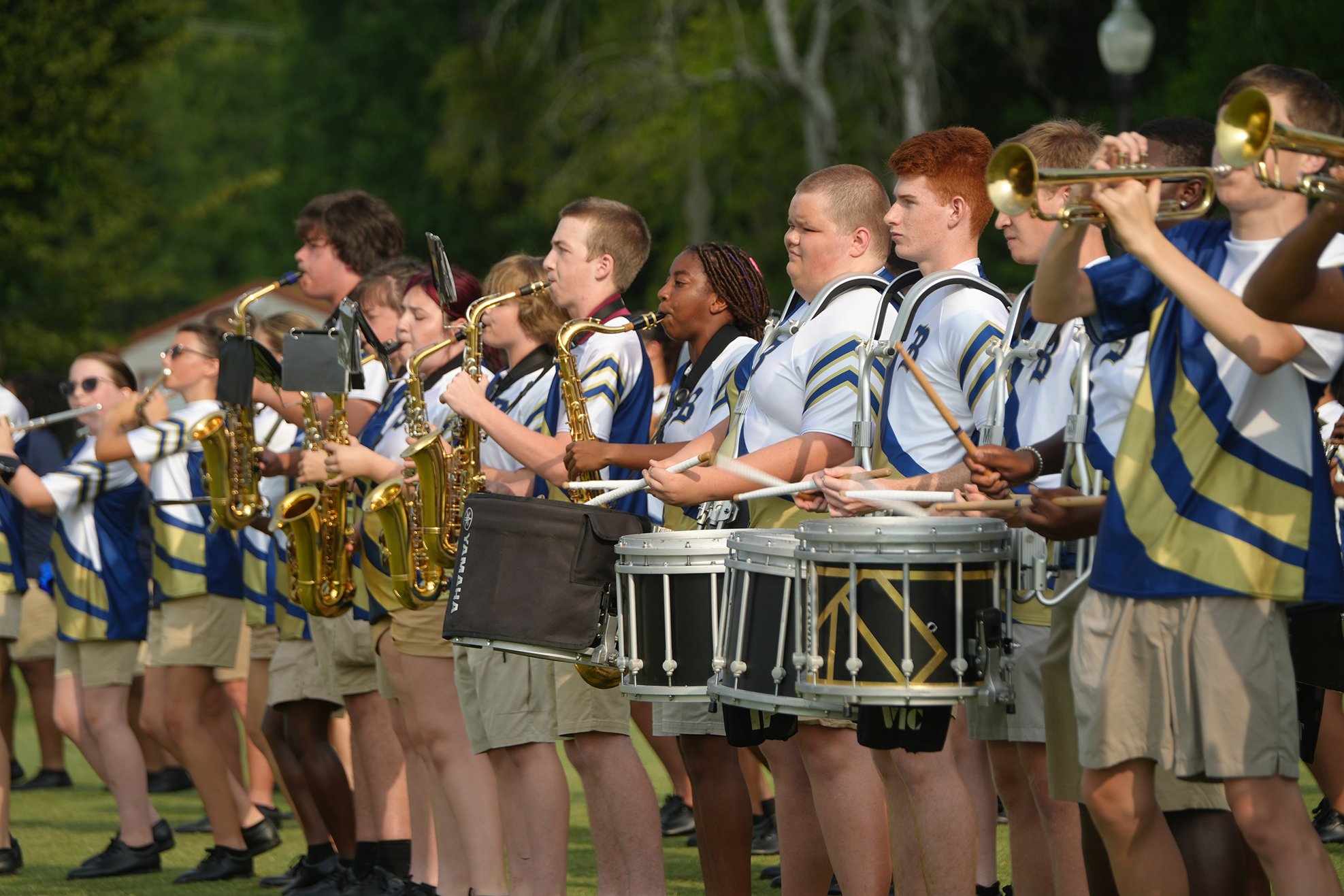 Band members, in blue, white, and yellow uniforms, holding instruments, and drums, on a grassy field.