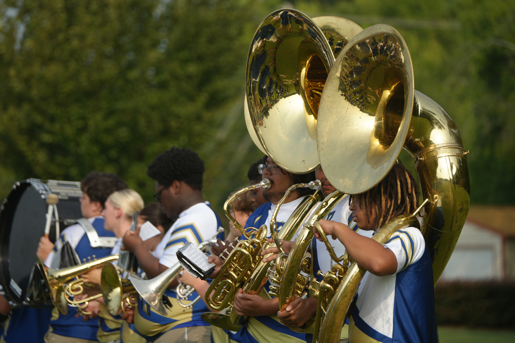 Several individuals in blue and white uniforms play brass instruments on a field with trees in the background.