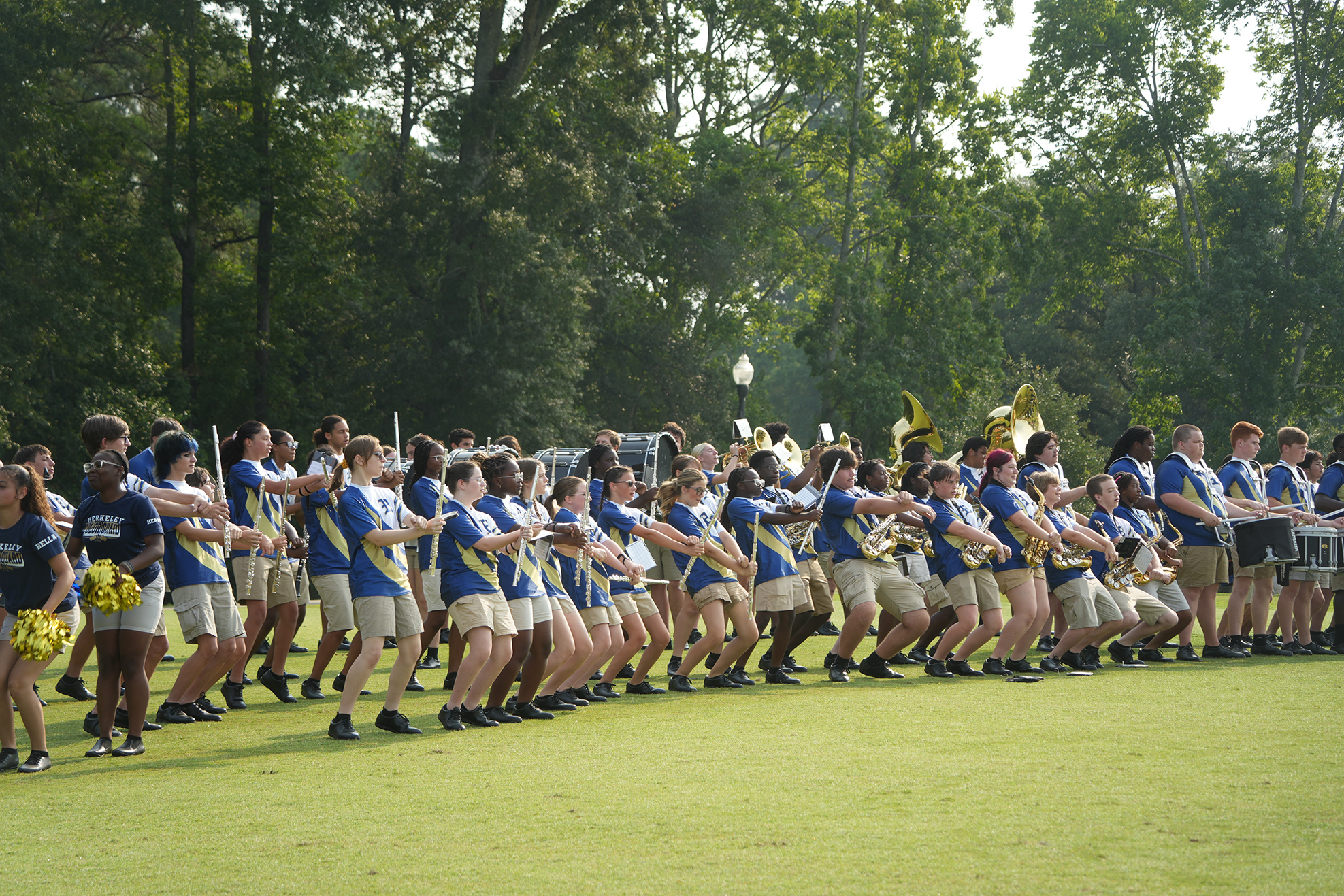 A marching band performs on a grassy field with trees in the background, wearing blue and white uniforms.