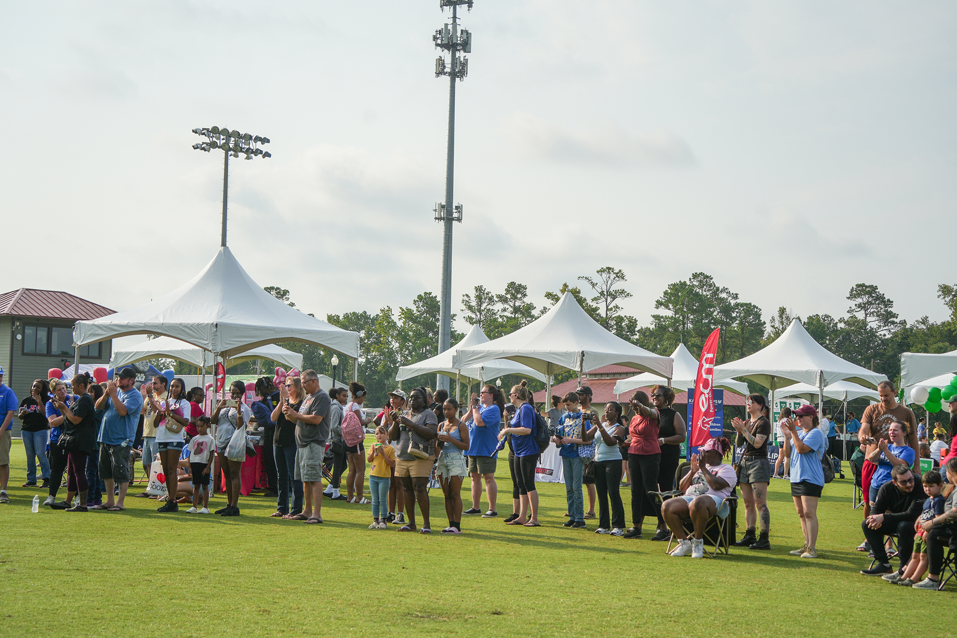 A large group of people stands on a field, some holding banners. Multiple tents are in the background.