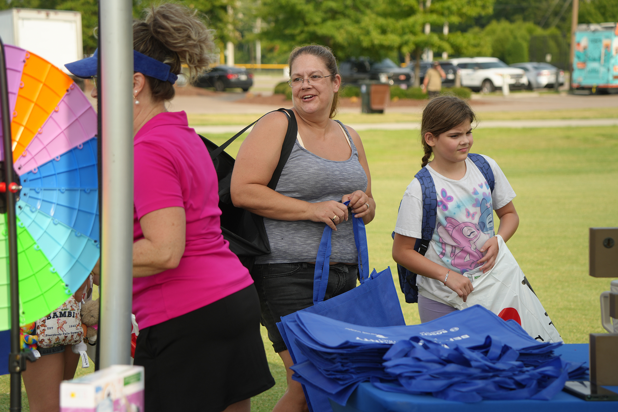 Three women stand outside by a table. One woman holds a colorful umbrella and a bag. Another holds blue bags. A young girl stands by the table.