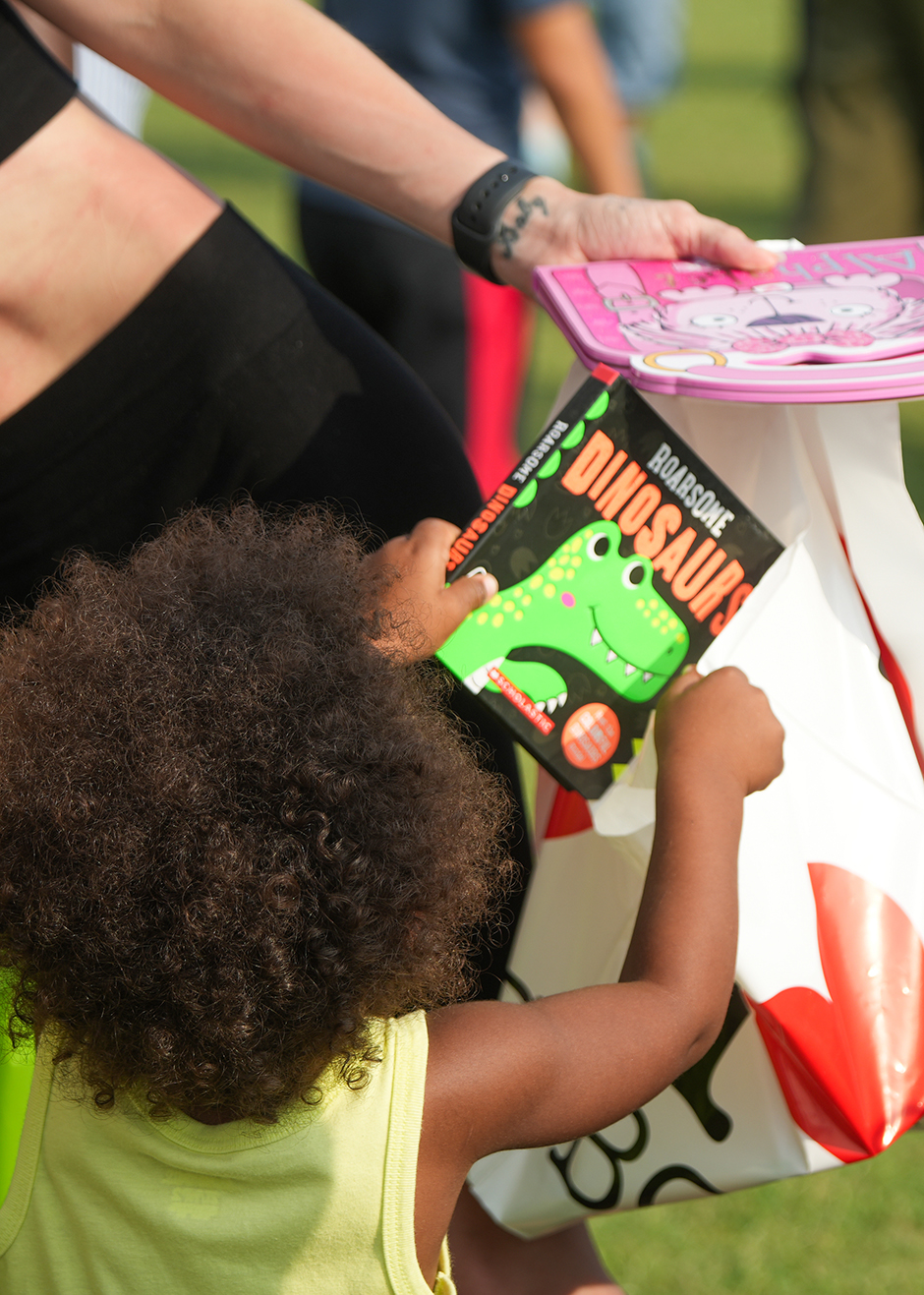 Child holding a dinosaur book in front of a bag with a red logo. Adult beside them holding a pink book.