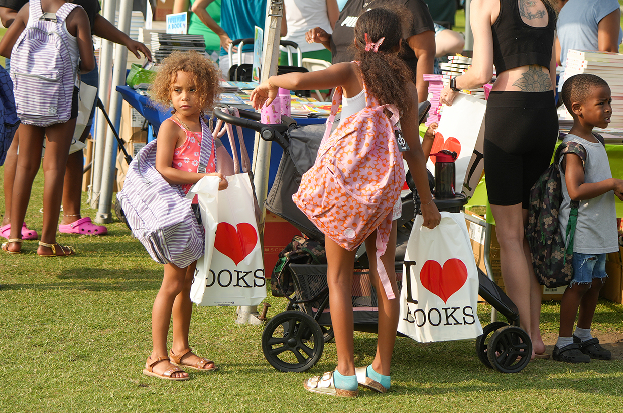 Children in casual clothing on grass, holding white bags with red hearts, some carrying backpacks.