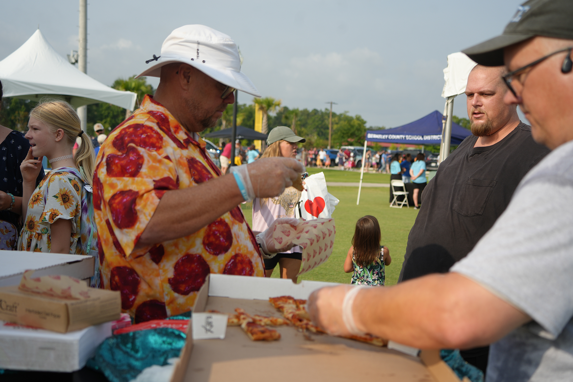 Three men in a food area with one man placing pizza on a tray, others watching.