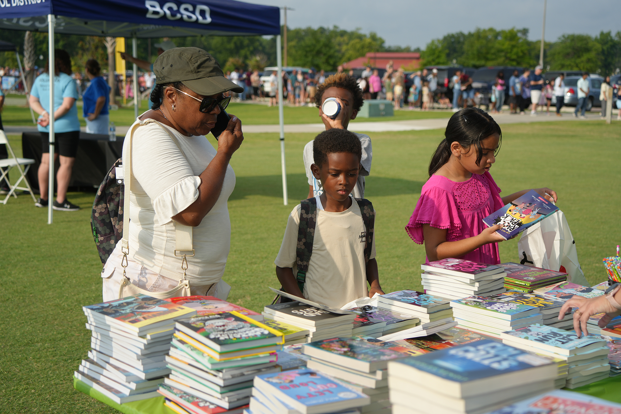 People are browsing a table with stacks of books. A woman is taking a photo of a young boy.