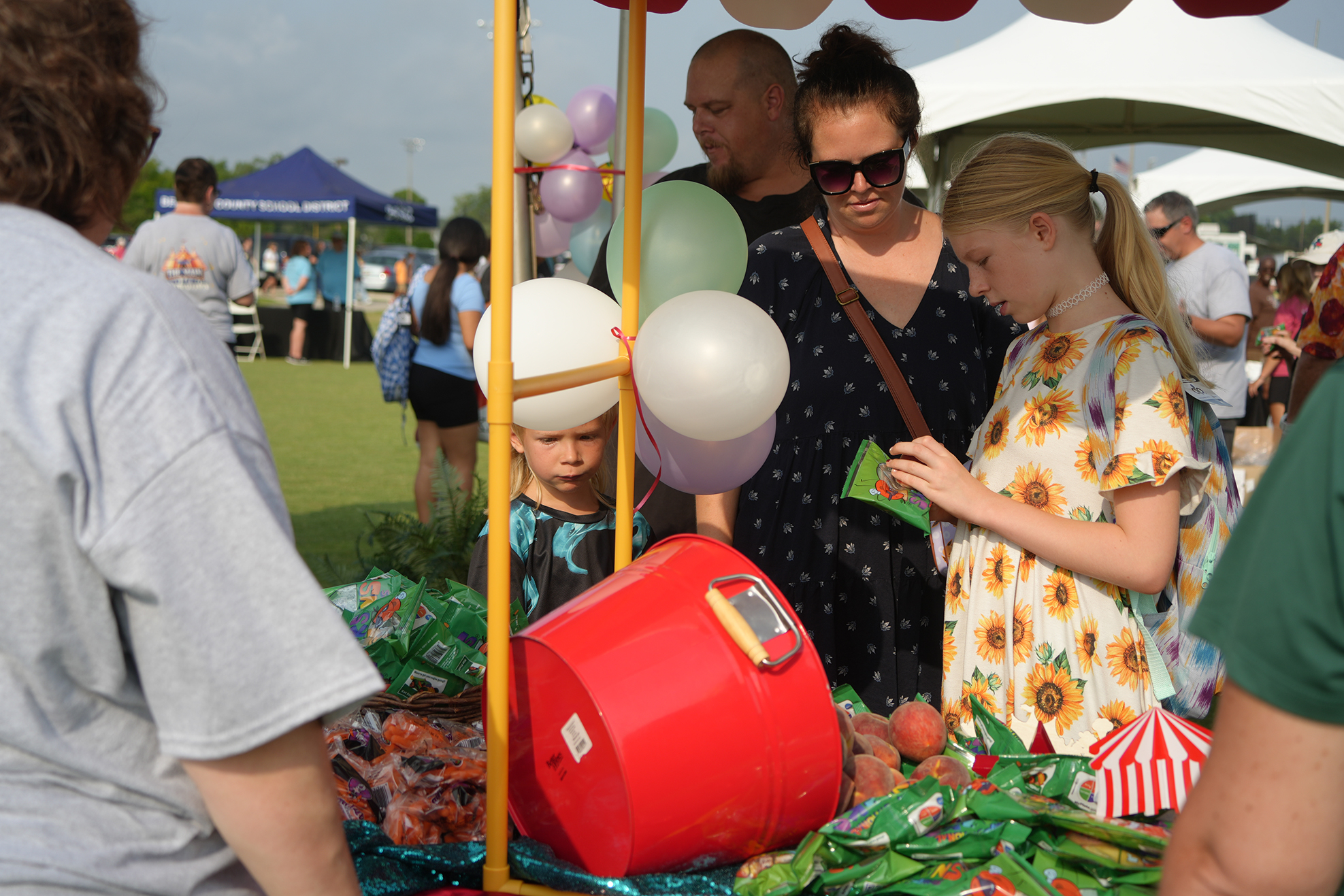 People gather around a table with a red bucket, balloons, and snacks. A girl examines items.