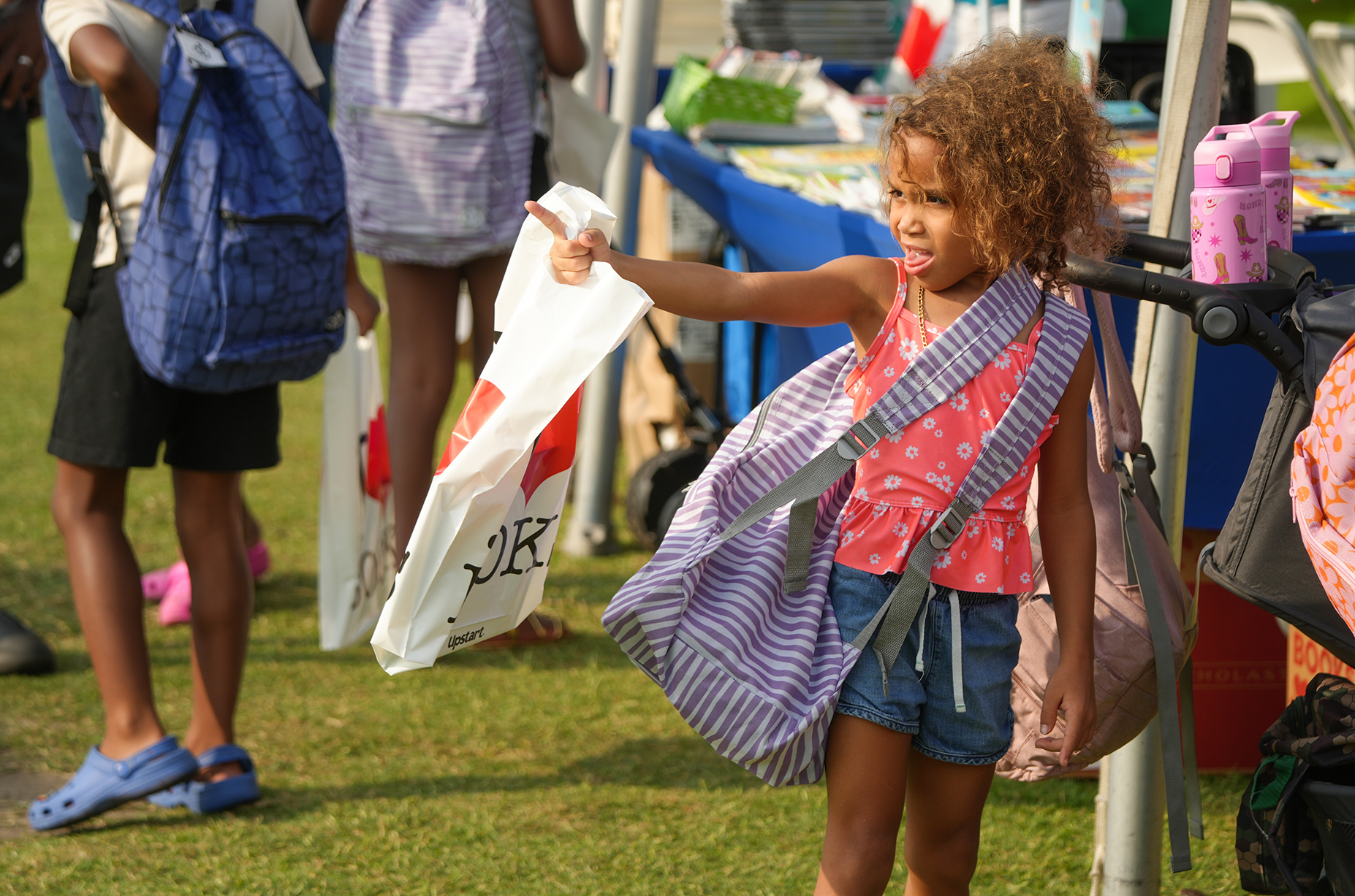 A young girl holds a white bag, wearing a pink top, denim shorts, and carrying a backpack.