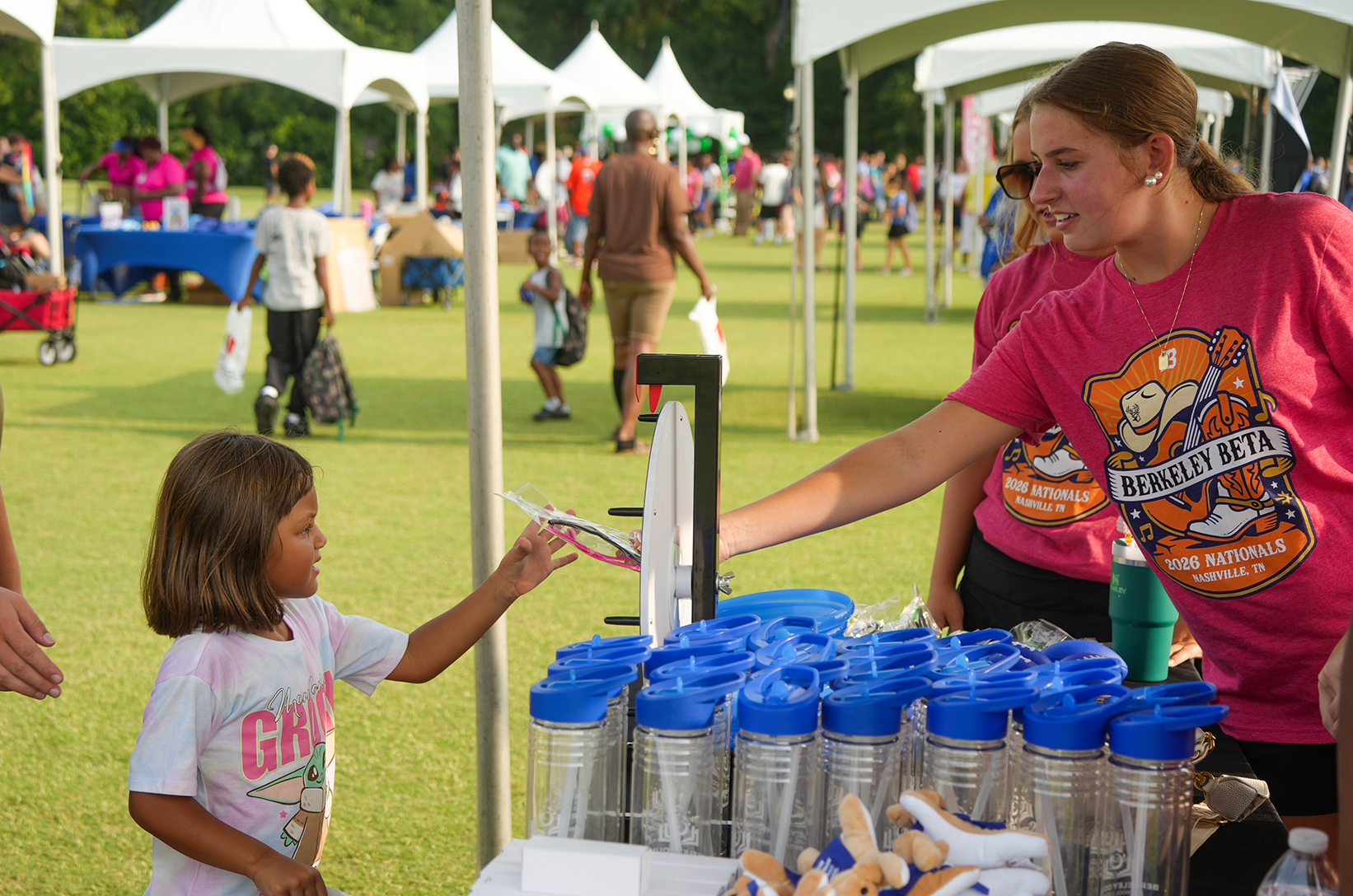 A young girl hands a paper to a woman at a booth with containers, surrounded by tents on a grassy field.