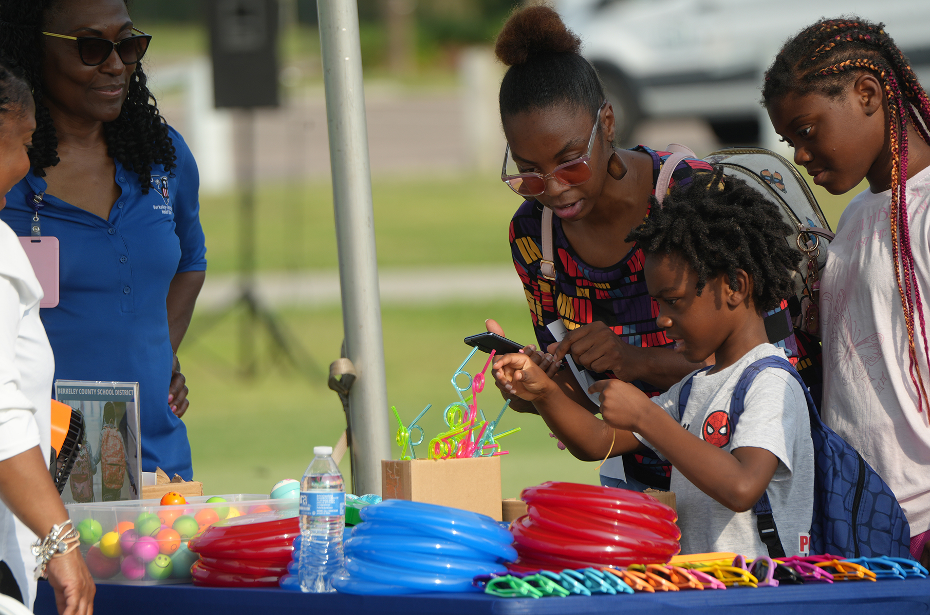 A woman and three children are at an outdoor table with toys. The child uses a cellphone.