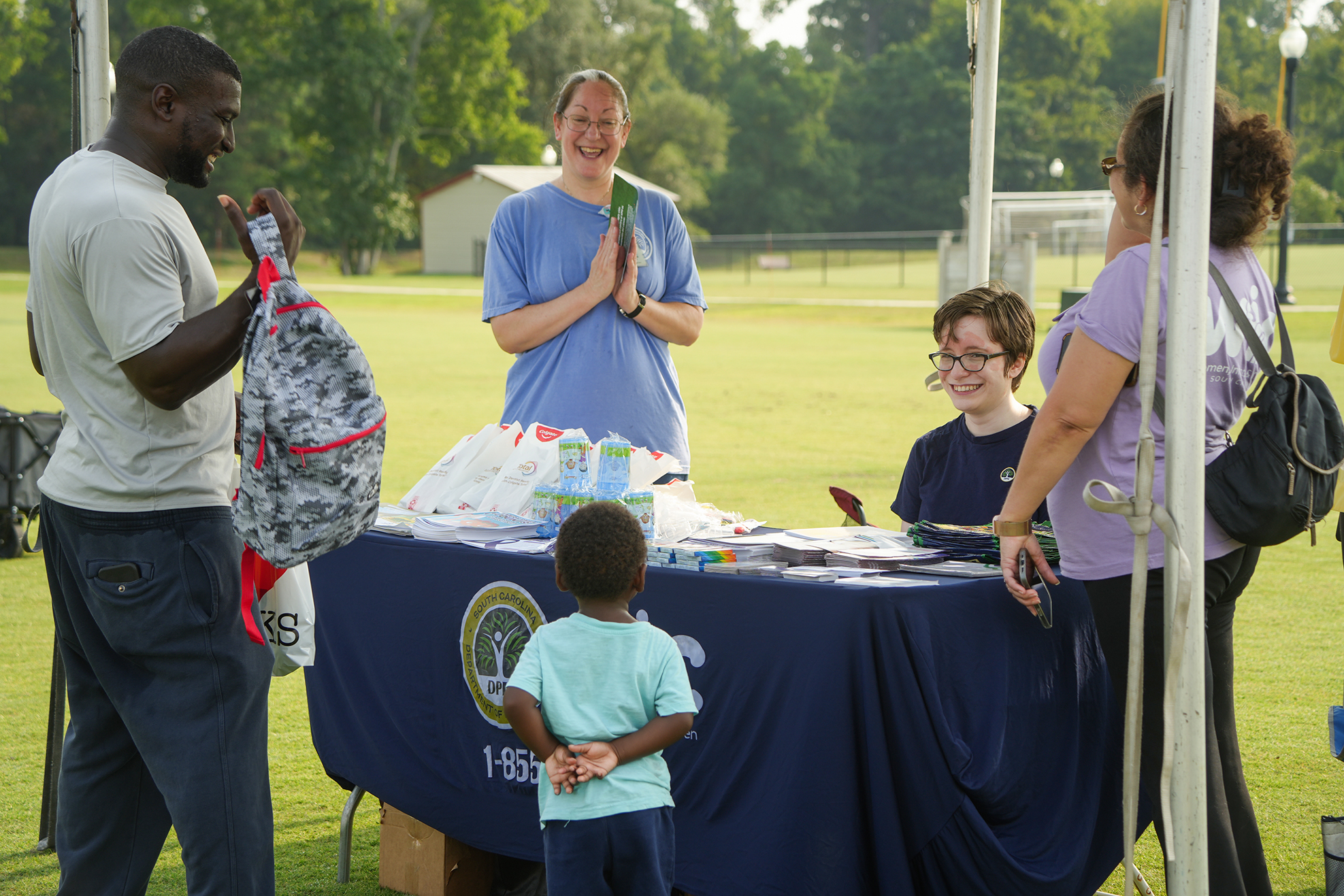 A group of people gather around a table. A man hands a backpack to a child. Another child sits at the table.