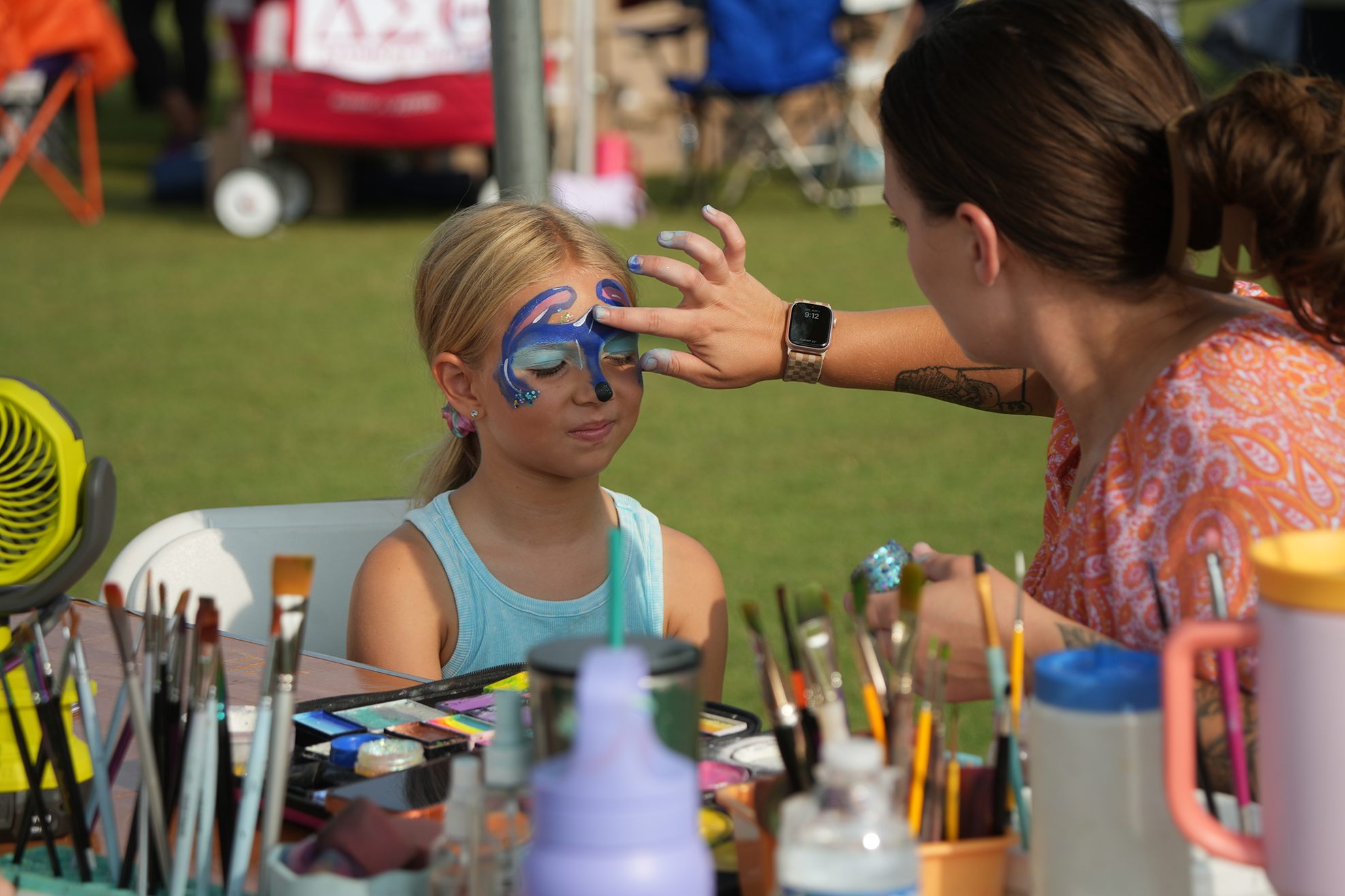 A woman applies blue paint to a young girl's face in an outdoor setting with tables and brushes.
