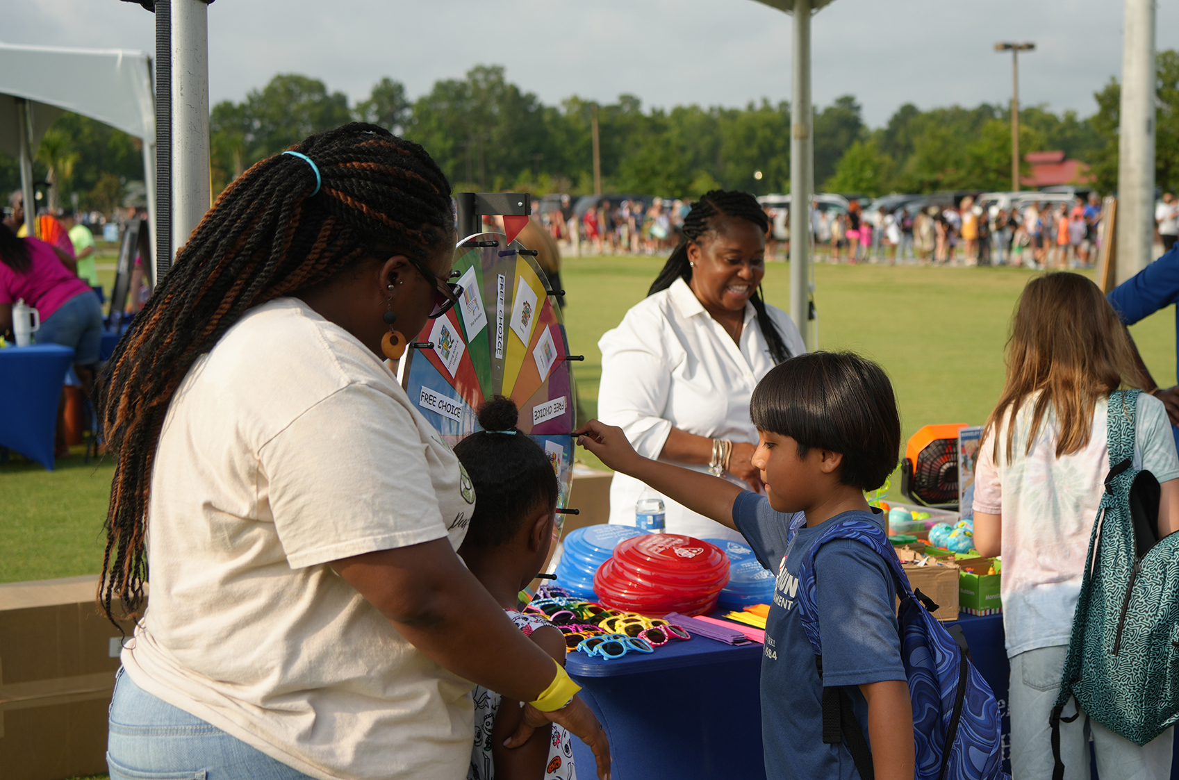 A woman in white and a child in a blue shirt interact at an outdoor event, with a table full of toys and other people in the background.