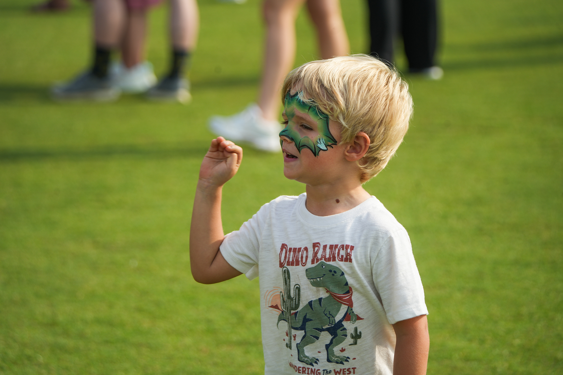 A child with a face painted green stands on a green lawn, wearing a dinosaur-themed white t-shirt.