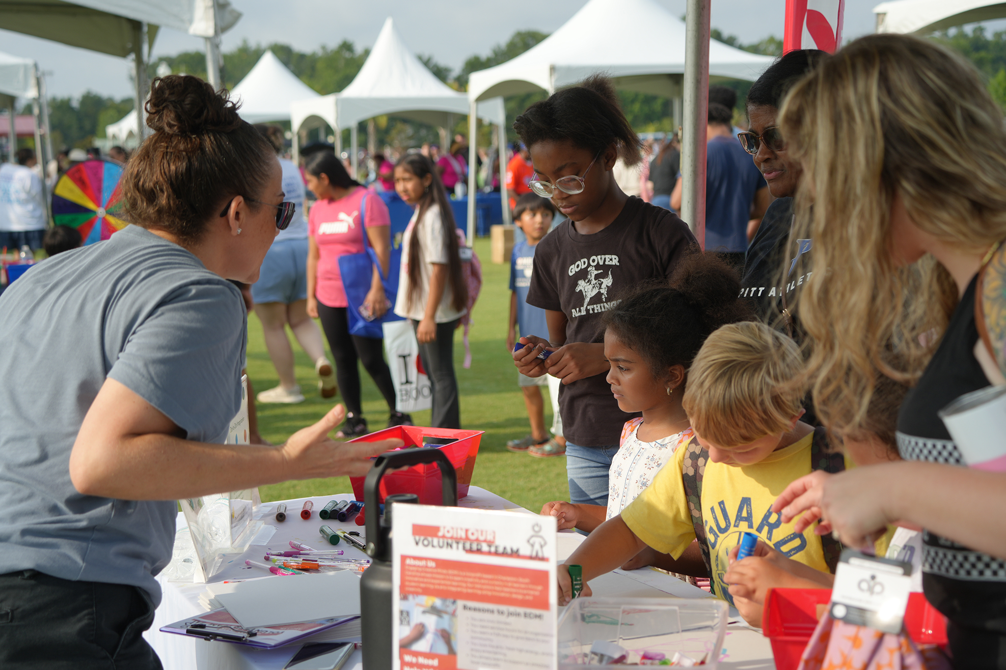People gather around a table with a woman in a gray shirt. Children sit, one with a yellow shirt.